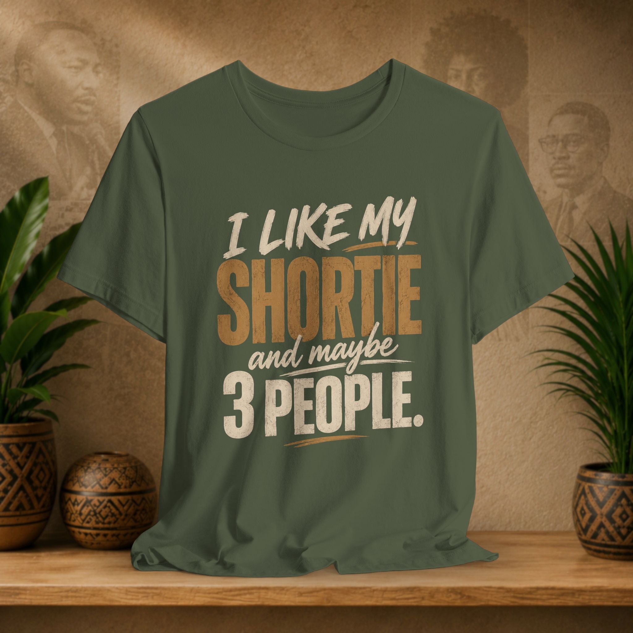 A military green graphic t-shirt displayed on a wooden shelf. The shirt features a humorous slogan printed in white and tan lettering: 'I LIKE MY SHORTIE and maybe 3 PEOPLE.