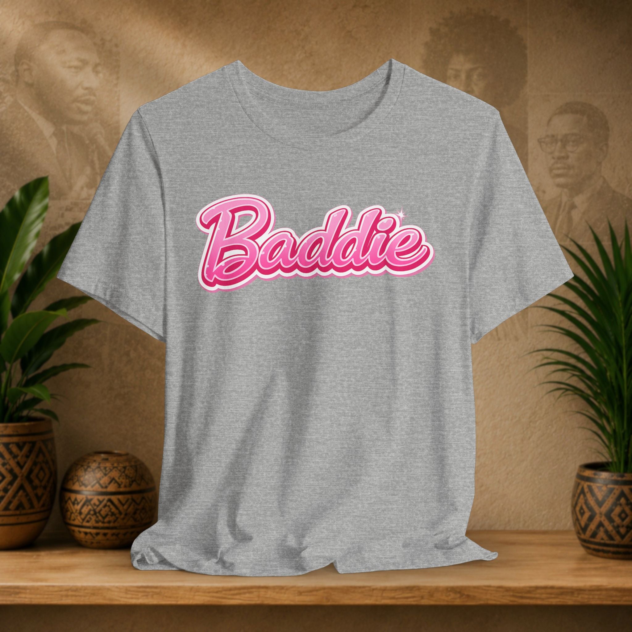 A heather gray short-sleeve t-shirt with 'Baddie' printed in pink cursive script with a white outline across the chest, displayed on a wooden shelf with potted plants and a textured background.