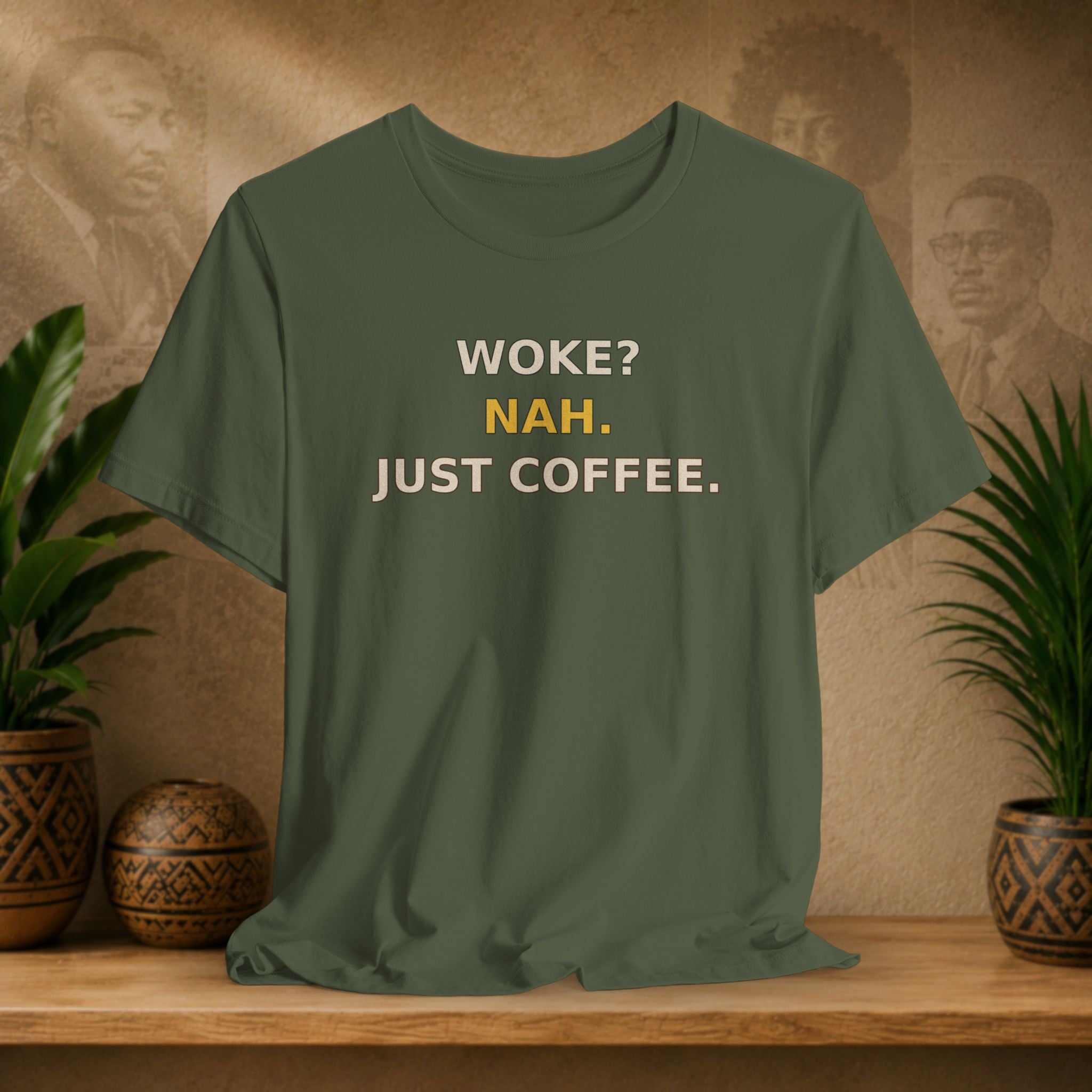 Green t-shirt with text 'WOKE? NAH. JUST COFFEE.' on a wooden surface with plants and a textured wall background.