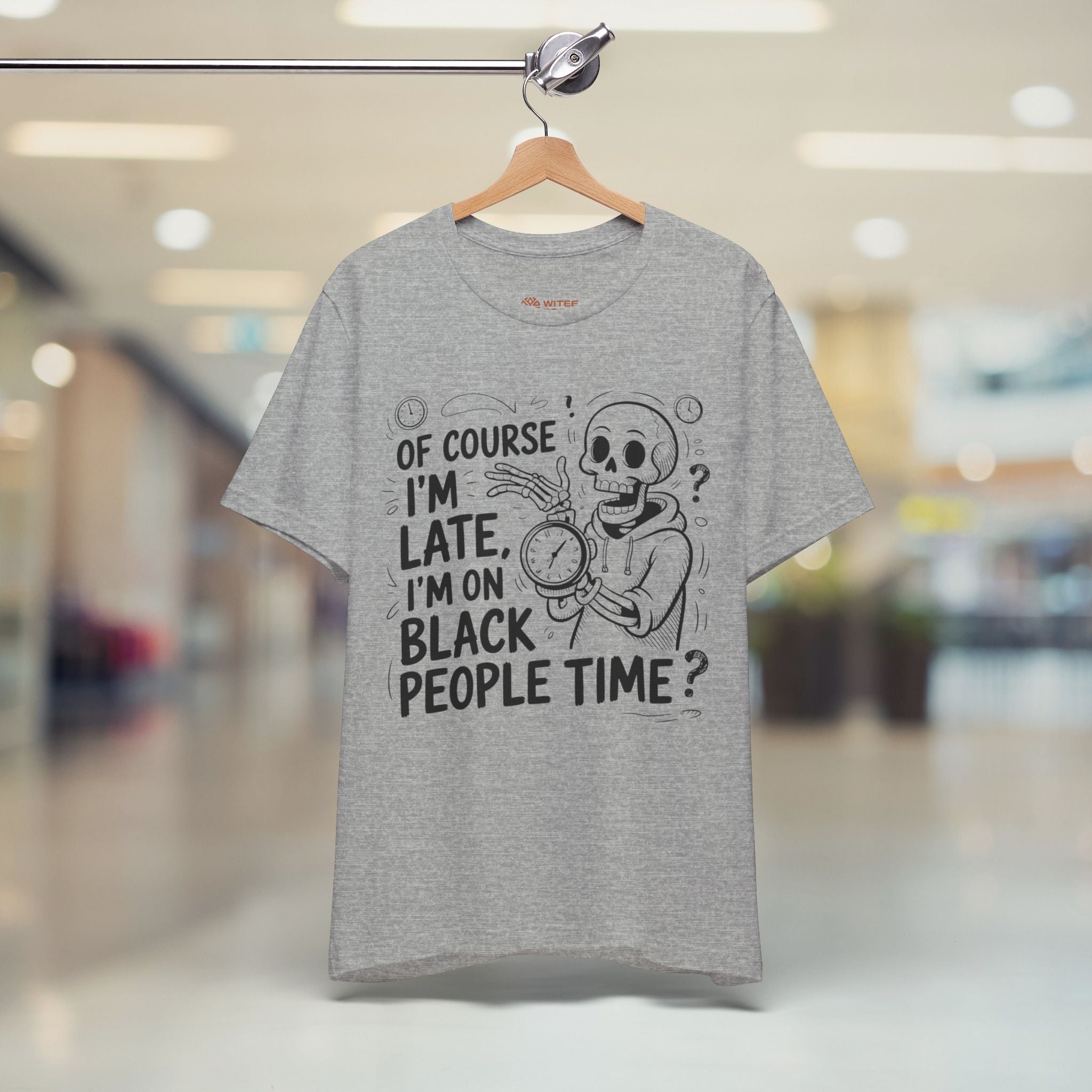 A heather gray graphic t-shirt hanging on a wooden hanger from a metal rack, set against a blurred indoor shopping mall background. The shirt features a black cartoon skeleton holding a clock with the text 'OF COURSE I'M LATE, I'M ON BLACK PEOPLE TIME?'.