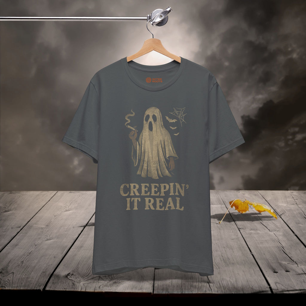 Gray t-shirt with ghost graphic and text 'Creepin' It Real' on a wooden surface with dark background