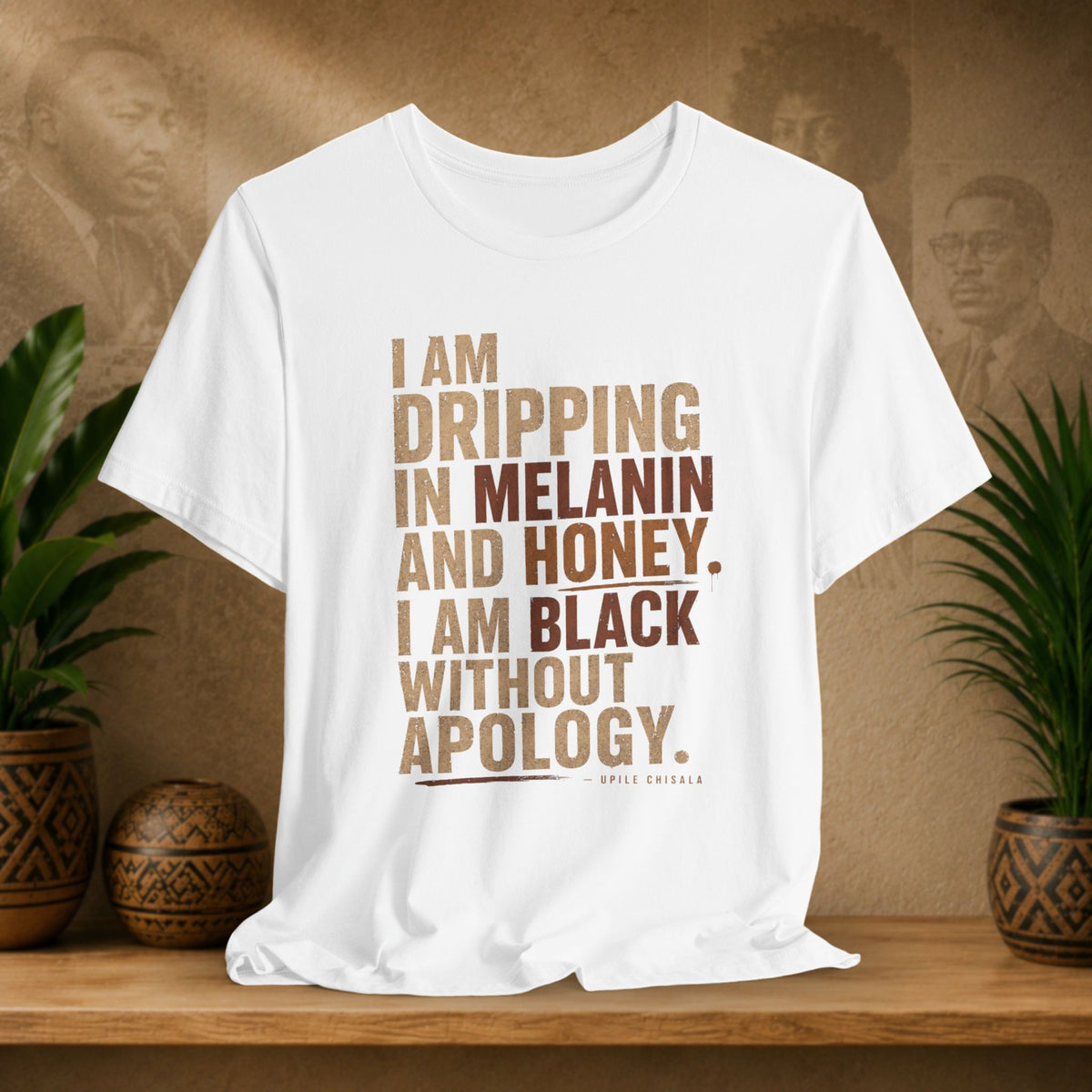 A white short-sleeve graphic T-shirt displayed on a wooden surface against a warm brown backdrop with decorative plants. The shirt features stacked text reading 'I AM DRIPPING IN MELANIN AND HONEY.