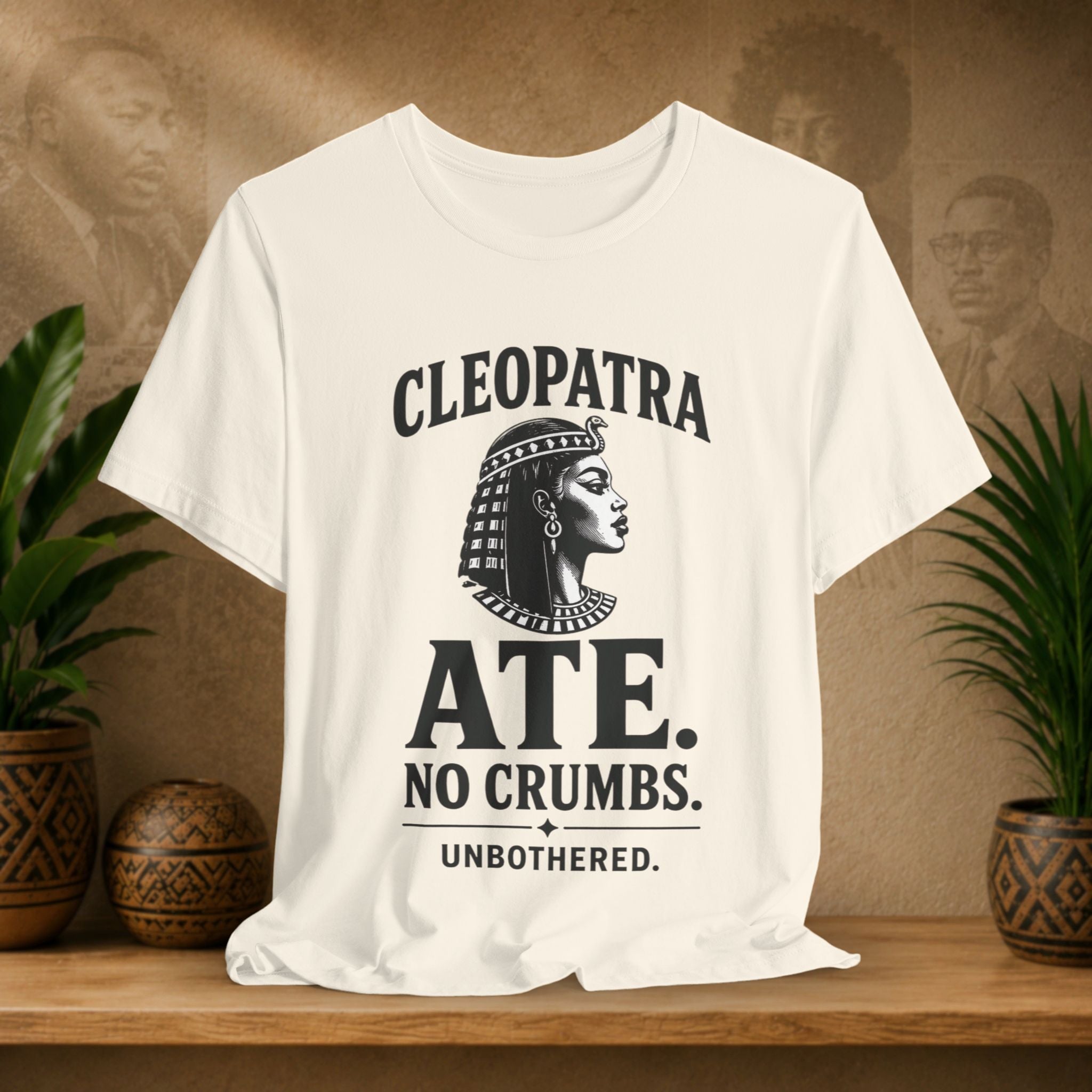 Beige t-shirt with Cleopatra graphic and text on a wooden surface with plants and decorative items.