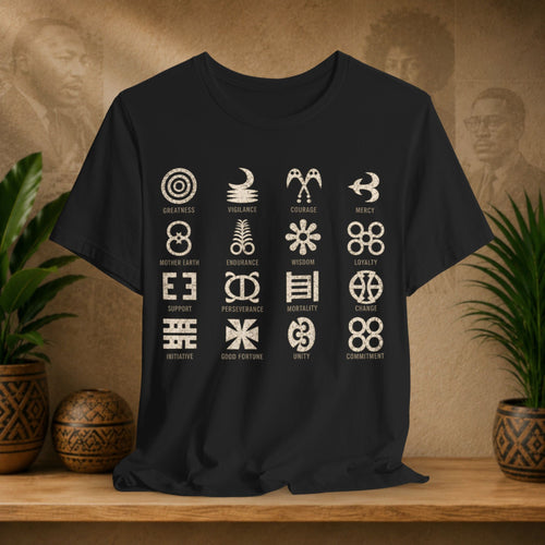 A black short-sleeve t-shirt with a grid of 16 Indigenous Australian symbols and their names printed in beige, displayed on a wooden shelf with decorative plants and pottery against a textured brown background.