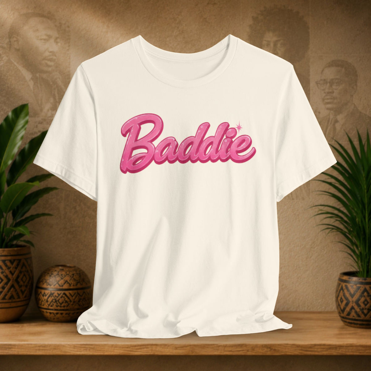 A beige short-sleeve T-shirt with the word 'Baddie' printed across the chest in a pink cursive font with a darker pink outline, displayed on a wooden surface with potted plants and a textured background.
