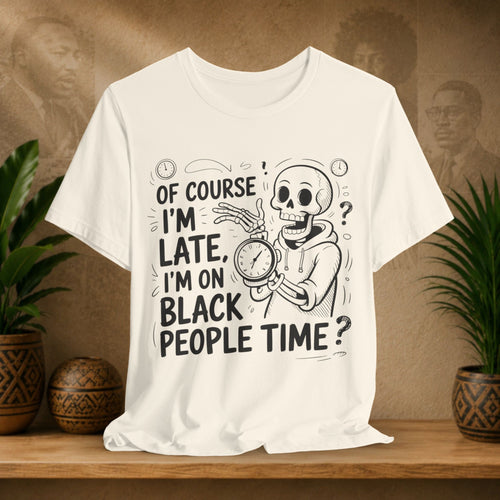 A beige short-sleeve graphic T-shirt displayed on a wooden surface, featuring a black cartoon skeleton holding a clock with the text 'OF COURSE I'M LATE, I'M ON BLACK PEOPLE TIME?'. The shirt is styled in a warm indoor setting with potted plants on both sides and a sepia-toned background.