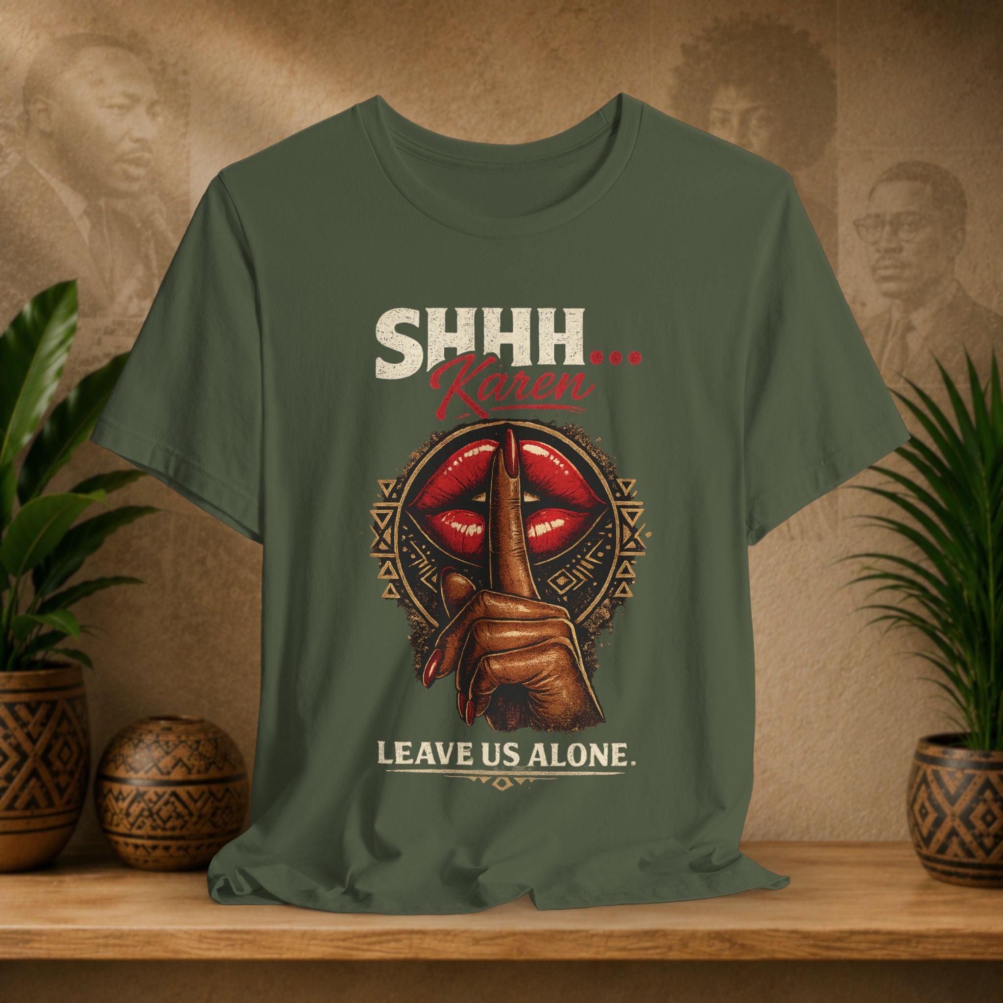 An olive green graphic t-shirt displayed on a wooden shelf with potted plants. The shirt features a graphic of a hand holding a red lips-and-moon symbol with the text 'SHHH.