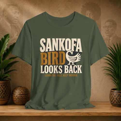 Green t-shirt with 'Sankofa Bird Looks Back' design on a wooden surface with plants and decorative items.