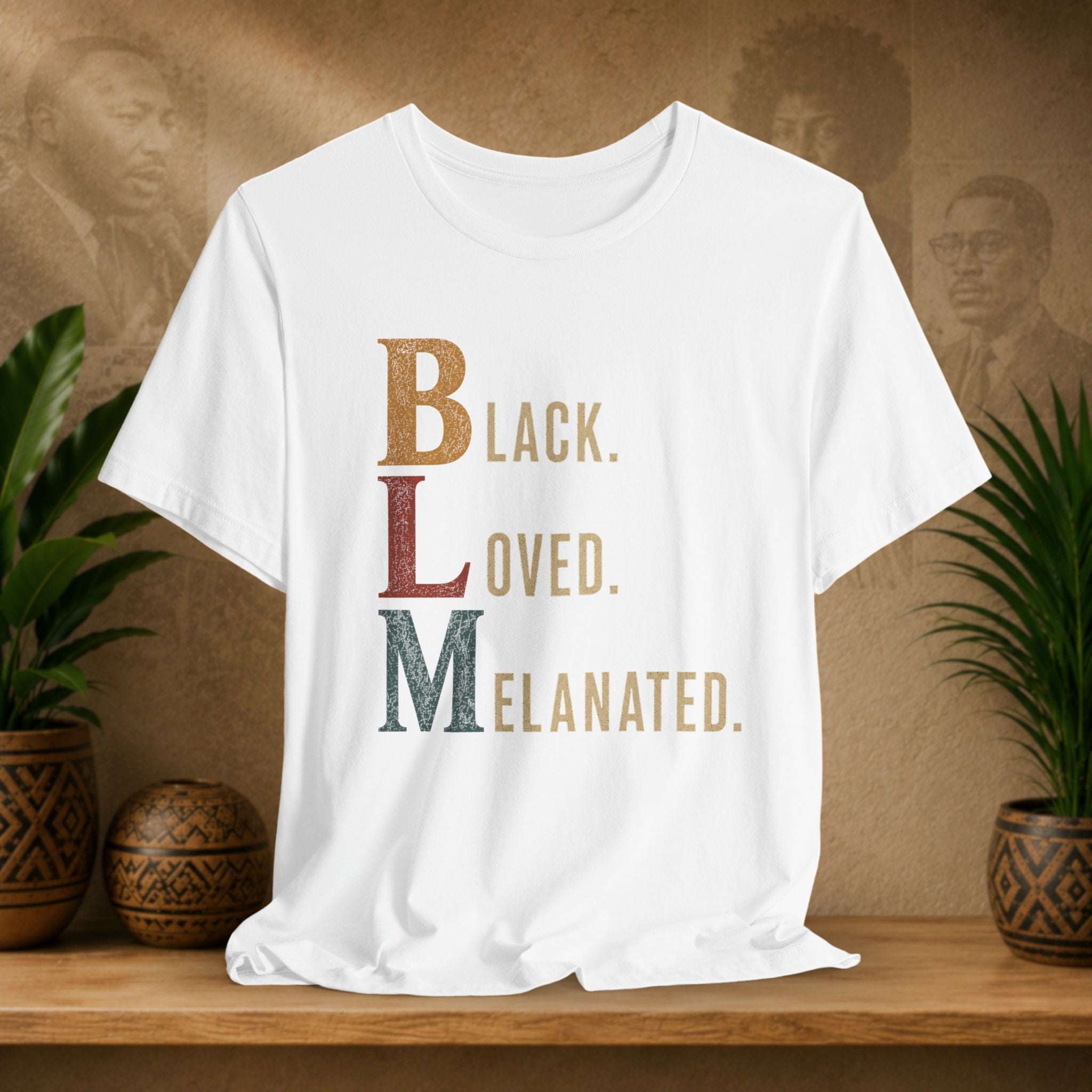 A white short-sleeve graphic T-shirt displayed on a wooden surface, featuring the text 'BLACK LOVED MELANATED' in stacked, stylized letters. The background is a warm brown wall with faint portraits, flanked by potted green plants.