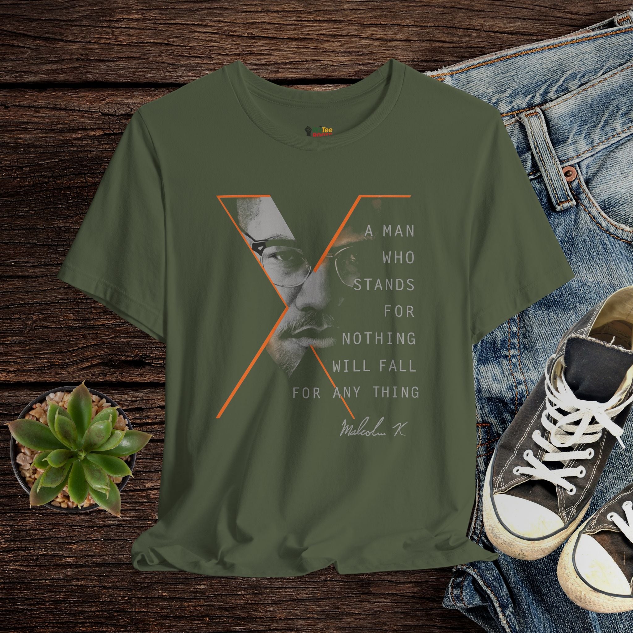 Green t-shirt with graphic design and text on a wooden surface with jeans and shoes.