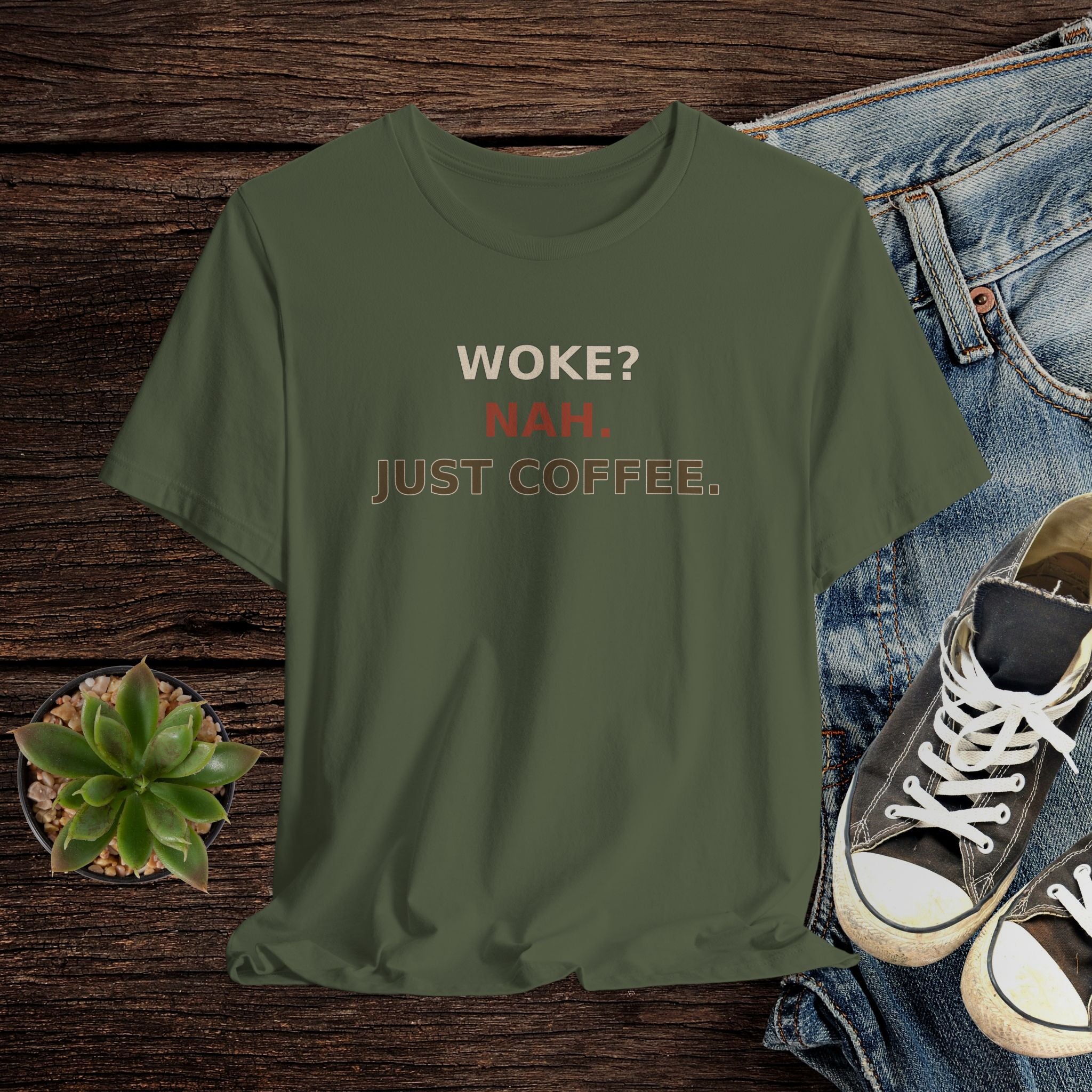 Green t-shirt with 'WOKE? NAH. JUST COFFEE.' text on a wooden surface with jeans and sneakers.