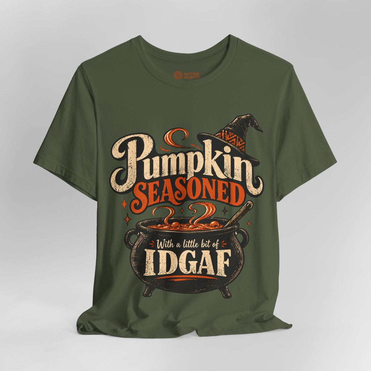 An olive green short-sleeve t-shirt with a Halloween graphic that reads 'Pumpkin Seasoned With a little bit of IDGAF'. The graphic features a witch hat, a cauldron, and orange and cream text, displayed on a neutral gray background.