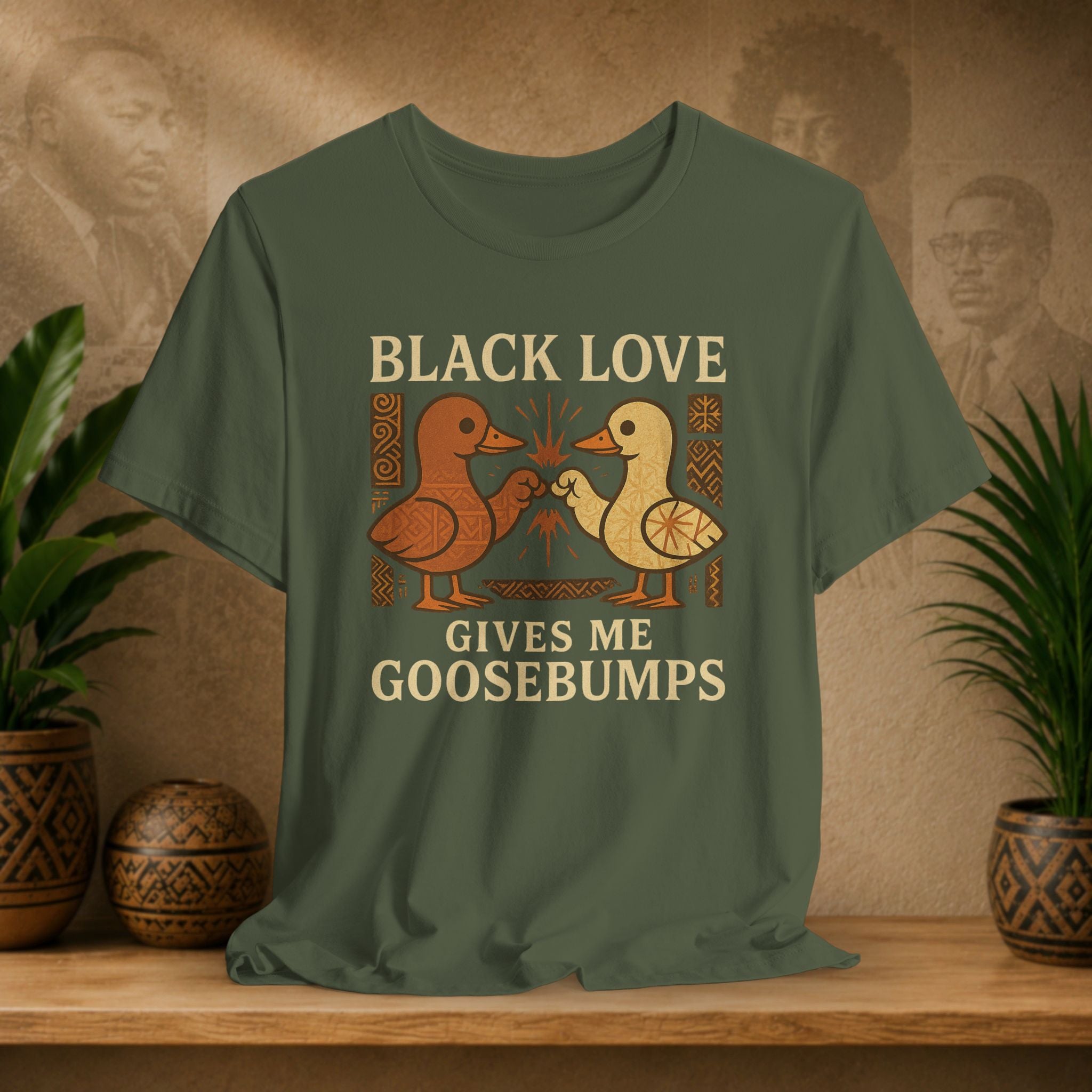Green t-shirt with 'Black Love Gives Me Goosebumps' text and duck graphics on a wooden surface with plants and wall art in the background.