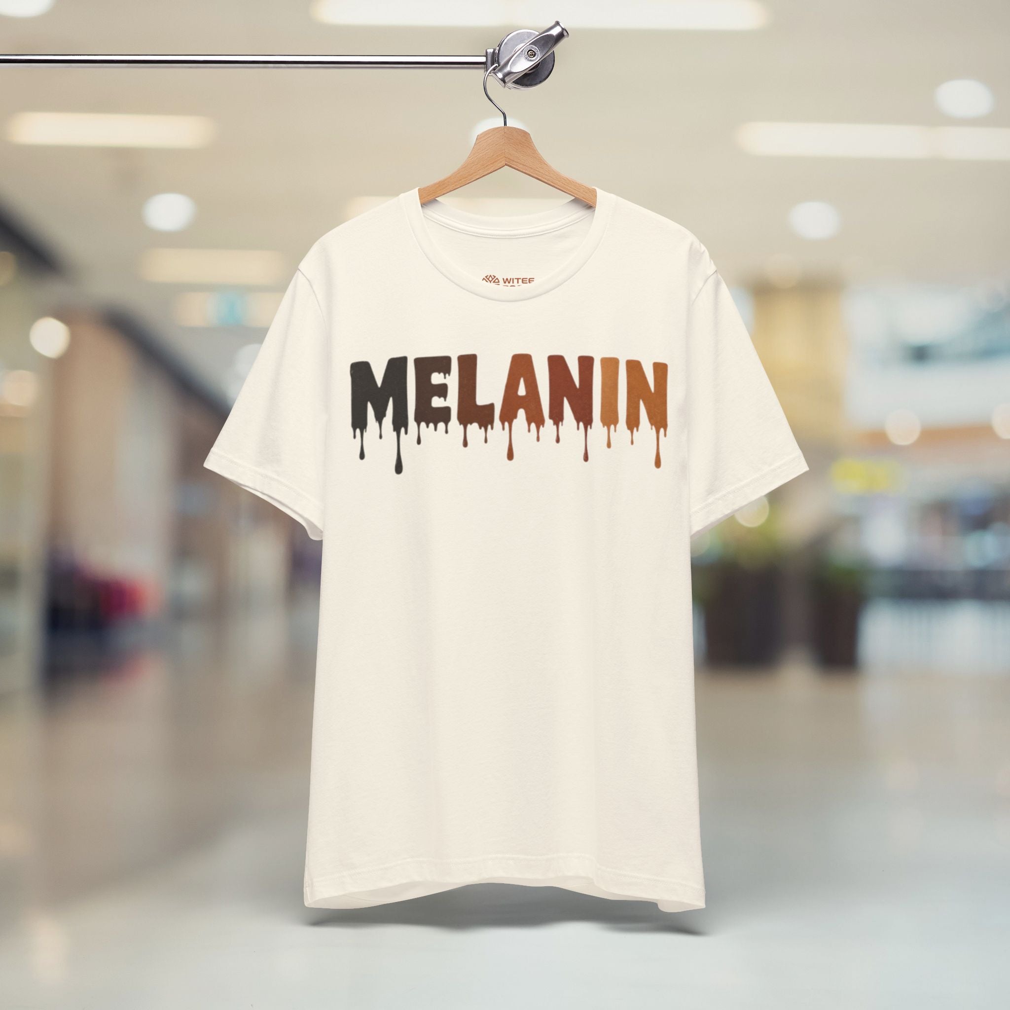 A beige short-sleeve t-shirt hanging on a wooden hanger from a clothing rack, featuring the word 'MELANIN' printed in a dripping paint style across the chest. The background is a blurred indoor shopping mall environment.