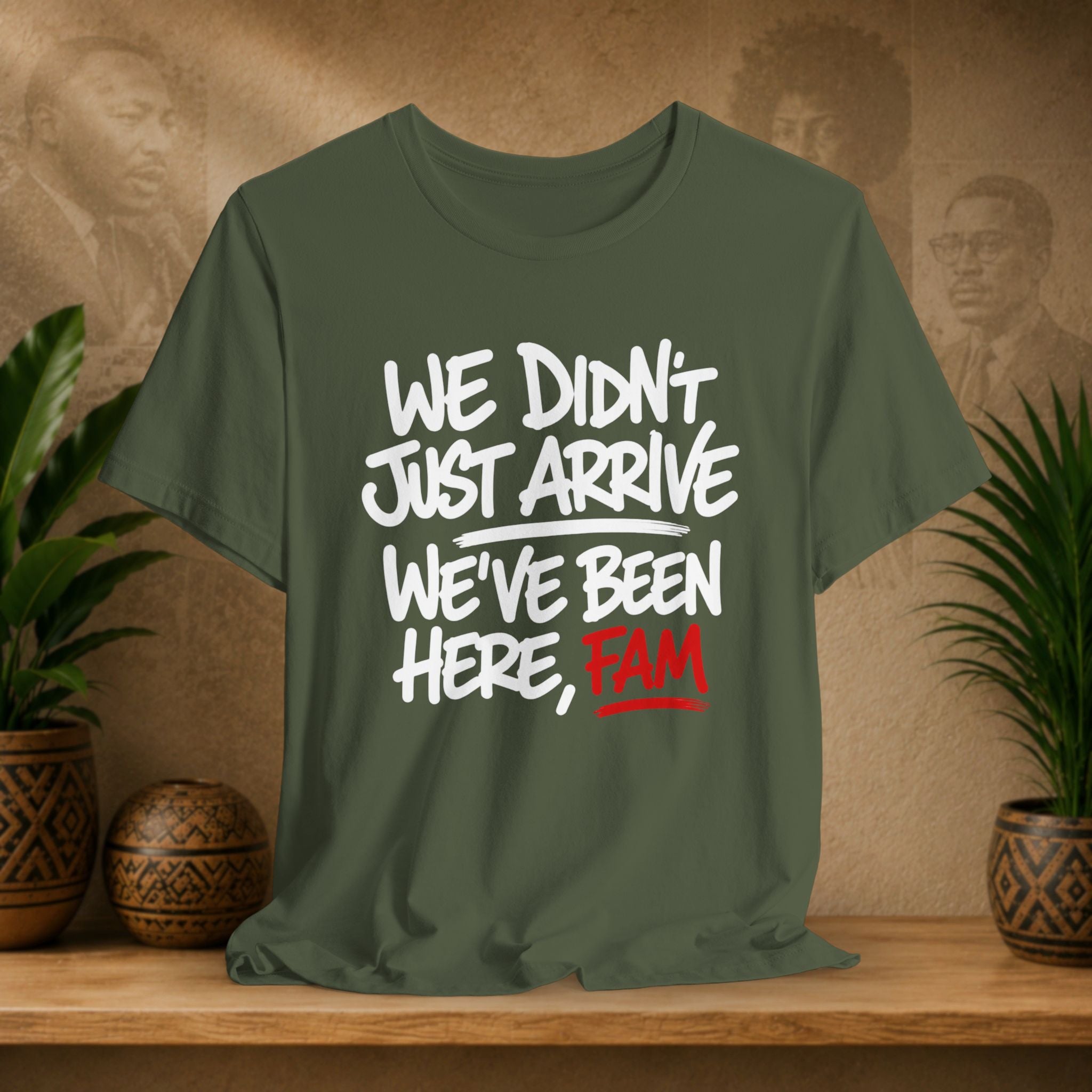 Green t-shirt with text 'WE DIDN'T JUST ARRIVE WE'VE BEEN HERE, FAM' on a wooden surface with plants and a wall with portraits in the background.