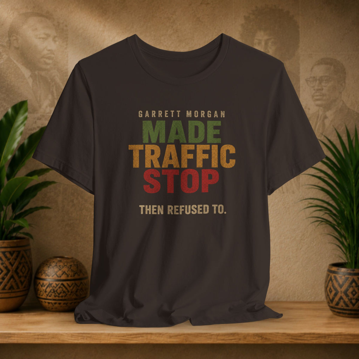 A brown graphic t-shirt displayed on a wooden shelf. The shirt features a retro-style text graphic that reads 'GARRETT MORGAN MADE TRAFFIC STOP THEN REFUSED TO.