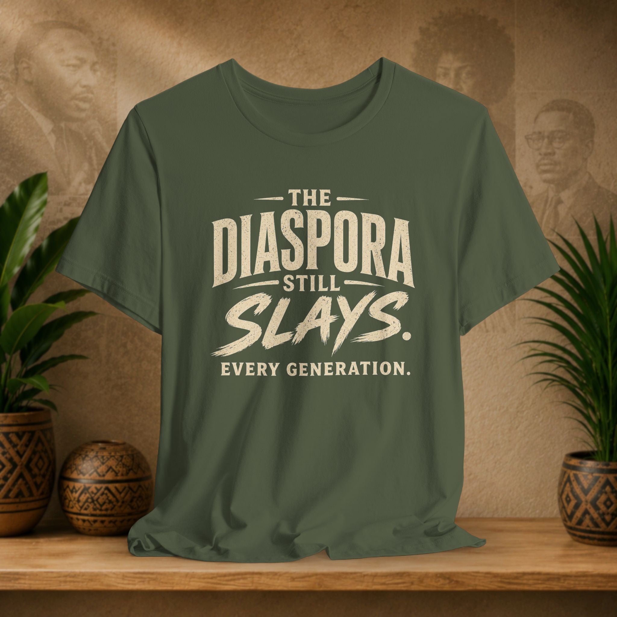 Green t-shirt with 'The Diaspora Still Slays. Every Generation.' text on a wooden surface with plants and decorative items.