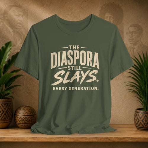 Green t-shirt with 'The Diaspora Still Slays. Every Generation.' text on a wooden surface with plants and decorative items.