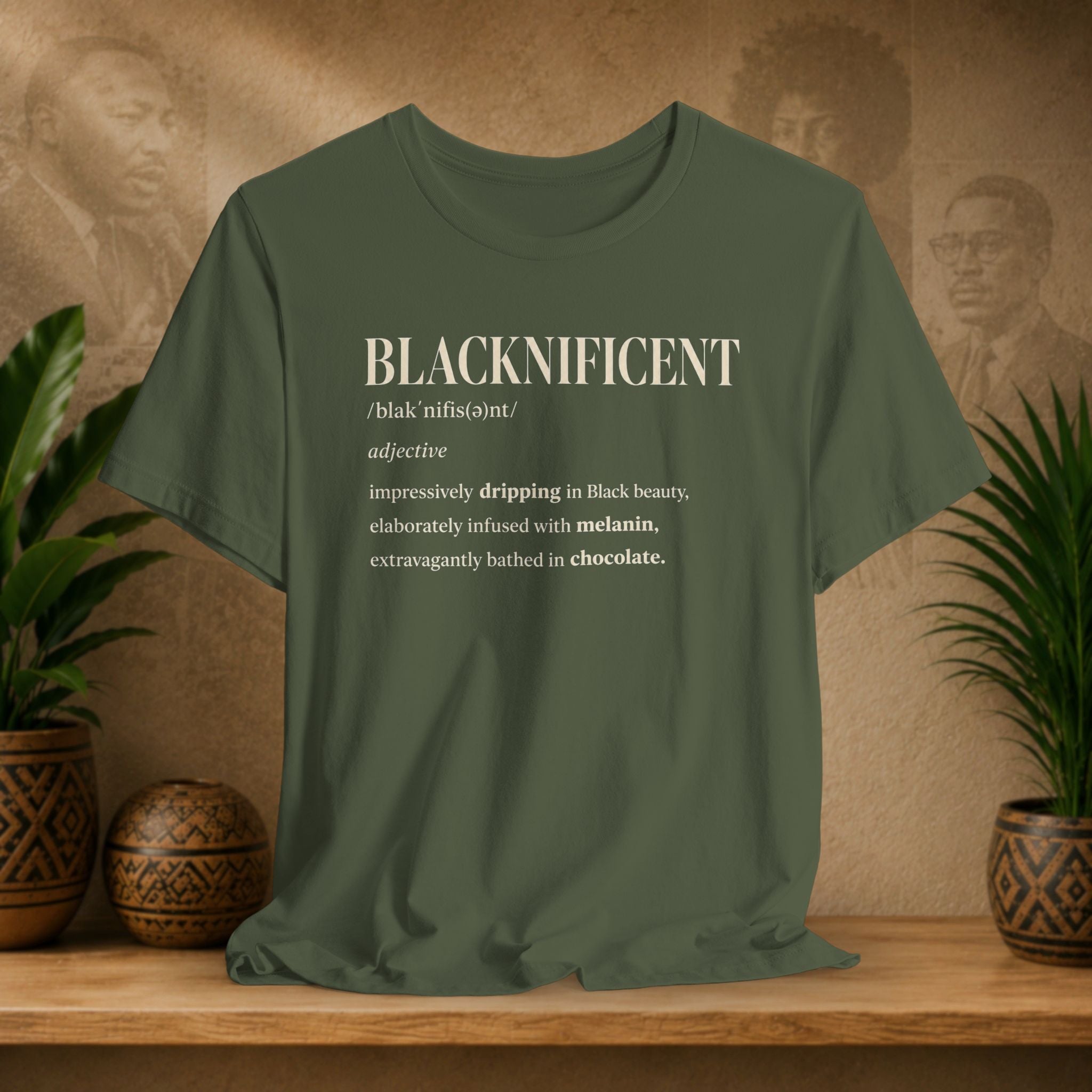 An olive green graphic t-shirt displayed on a wooden shelf with potted plants. The shirt features a white text graphic on the chest defining the word 'BLACKNIFICENT'.