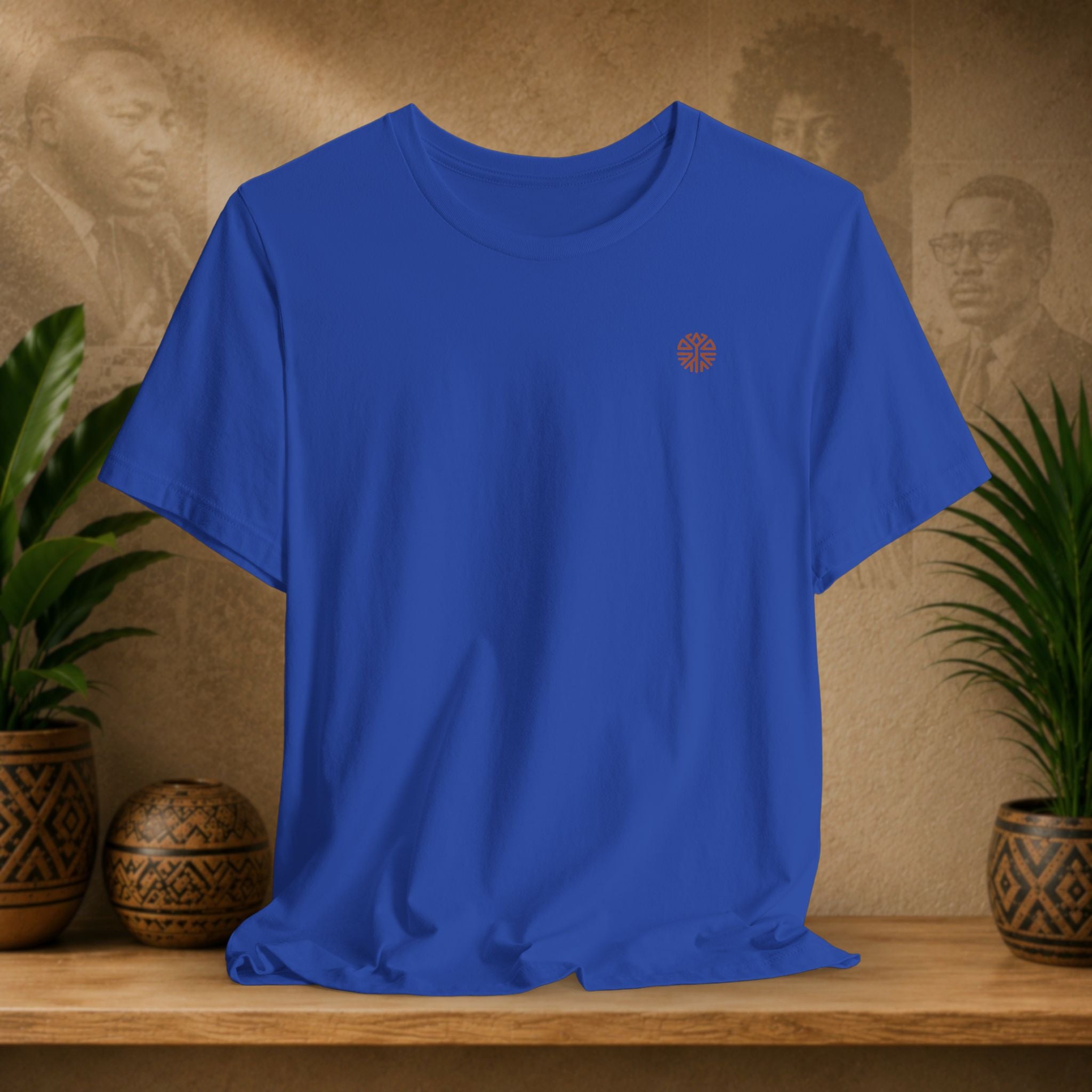 Blue t-shirt with a small red logo on a wooden surface with plants and a textured wall background