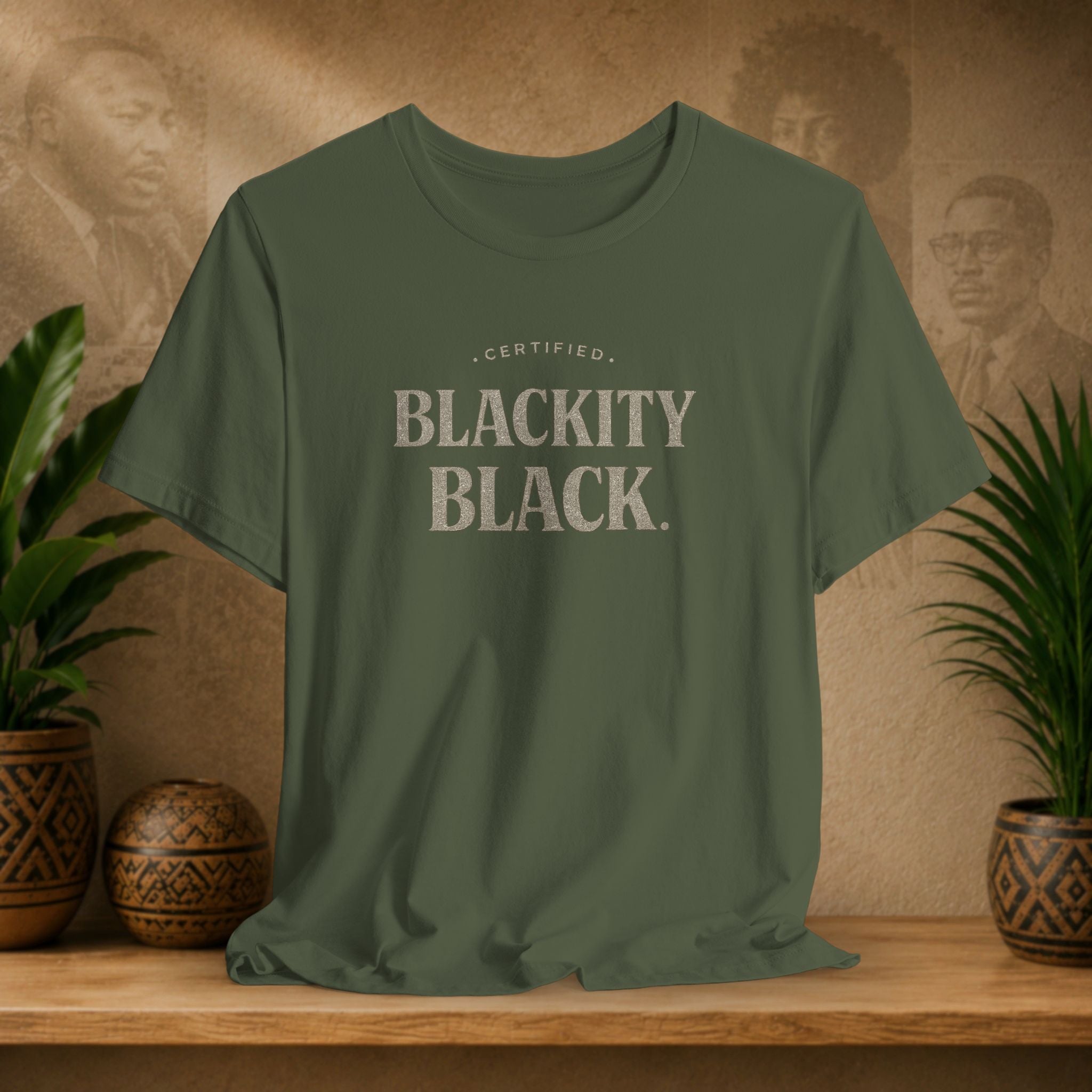 Green t-shirt with 'CERTIFIED BLACKITY BLACK' text on a wooden surface with decorative items and a background of portraits.
