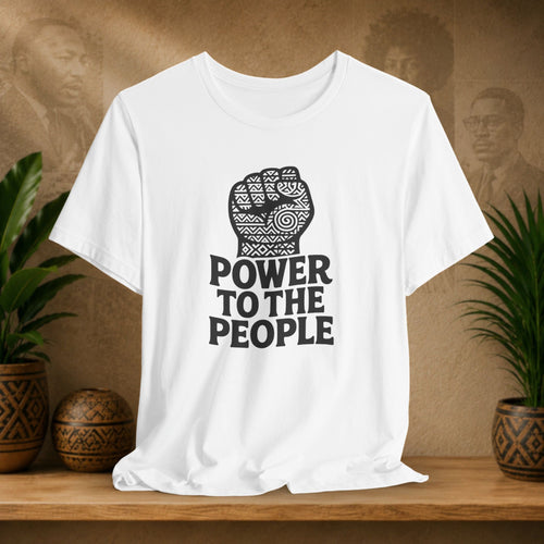 White t-shirt with 'Power to the People' design on a wooden surface with plants and a textured wall background.