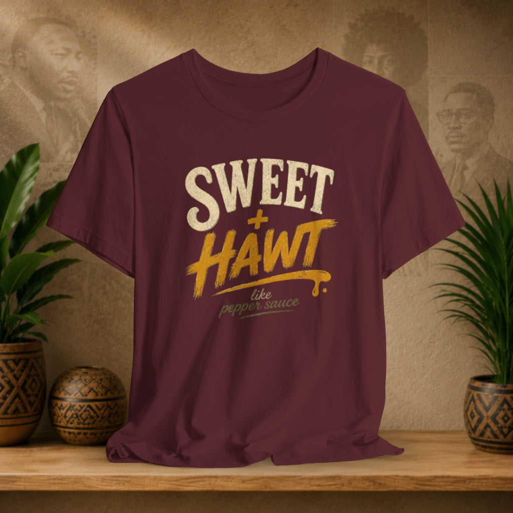 A maroon short-sleeve graphic T-shirt displayed on a wooden surface, with a warm beige wall and potted plants in the background. The shirt features a distressed graphic reading 'SWEET + HAWT like pepper sauce' in cream and yellow lettering.