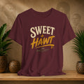 A maroon short-sleeve graphic T-shirt displayed on a wooden surface, with a warm beige wall and potted plants in the background. The shirt features a distressed graphic reading 'SWEET + HAWT like pepper sauce' in cream and yellow lettering.