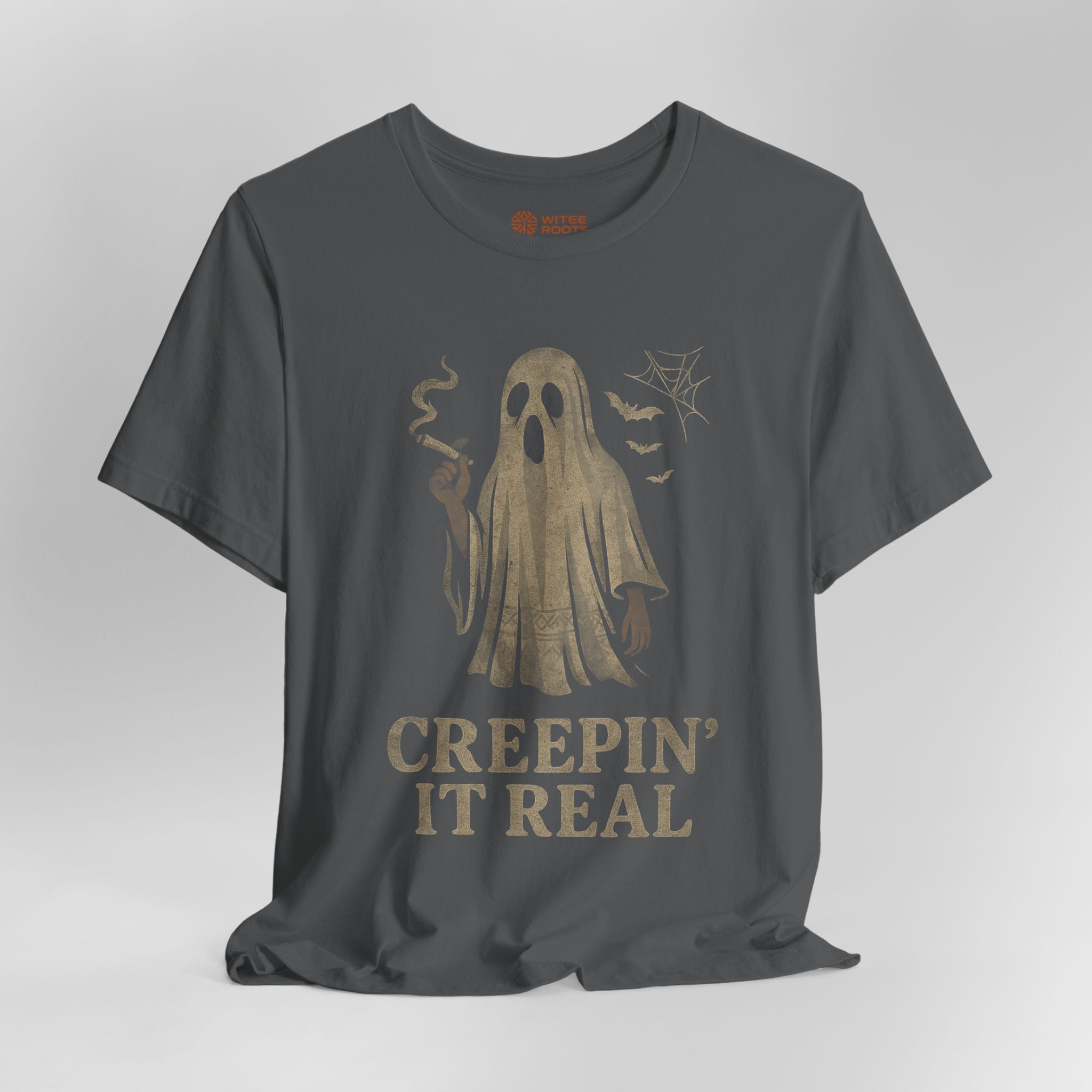 A dark charcoal gray t-shirt with a distressed beige graphic of a smoking ghost, bats, and spiderwebs, with the text 'CREEPIN' IT REAL' printed below.
