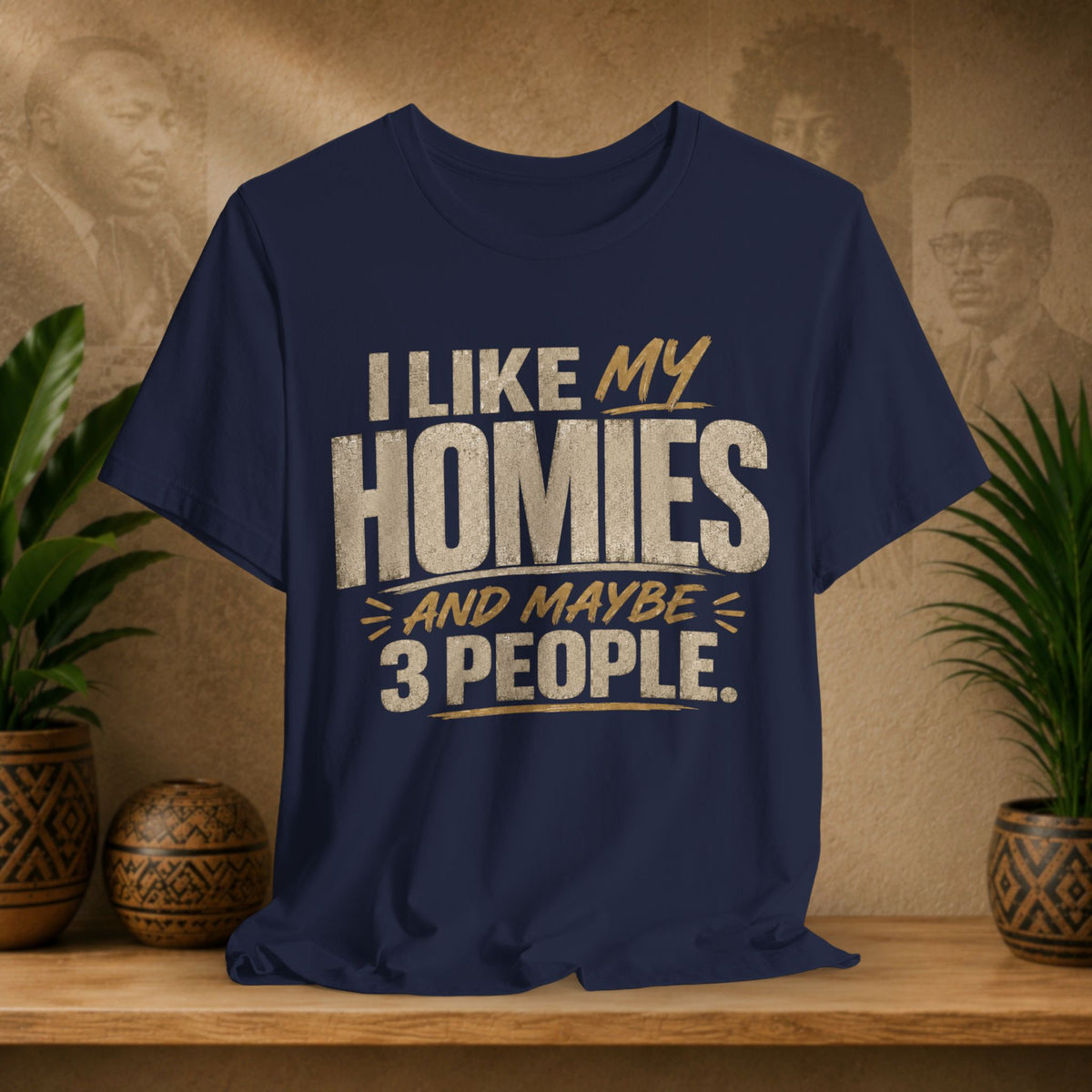 A navy graphic t-shirt displayed on a wooden shelf. The shirt features a humorous slogan printed in white and tan lettering: 'I LIKE MY HOMIES and maybe 3 PEOPLE.
