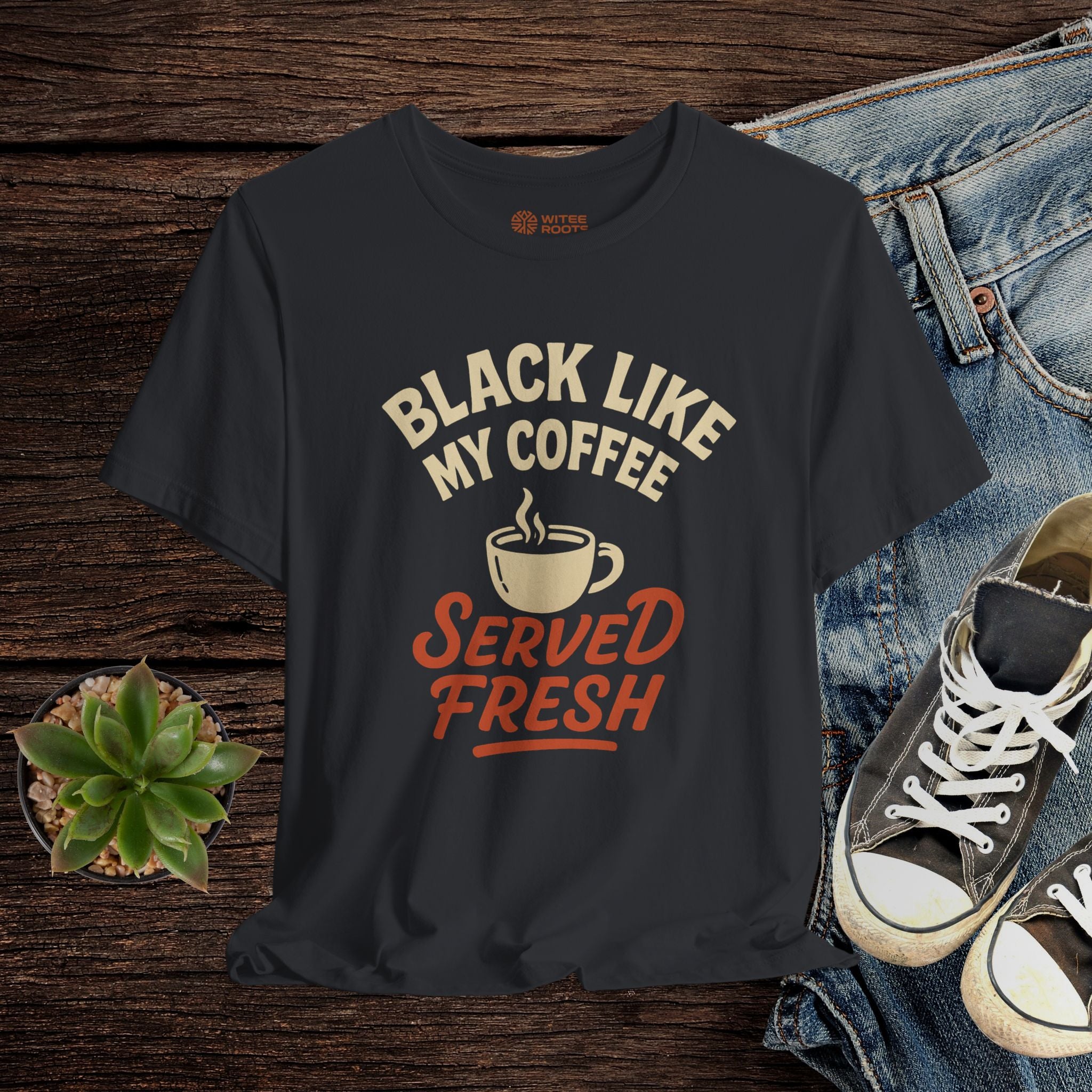 Black t-shirt with 'Black Like My Coffee Served Fresh' text and graphics on a wooden surface with jeans and sneakers.