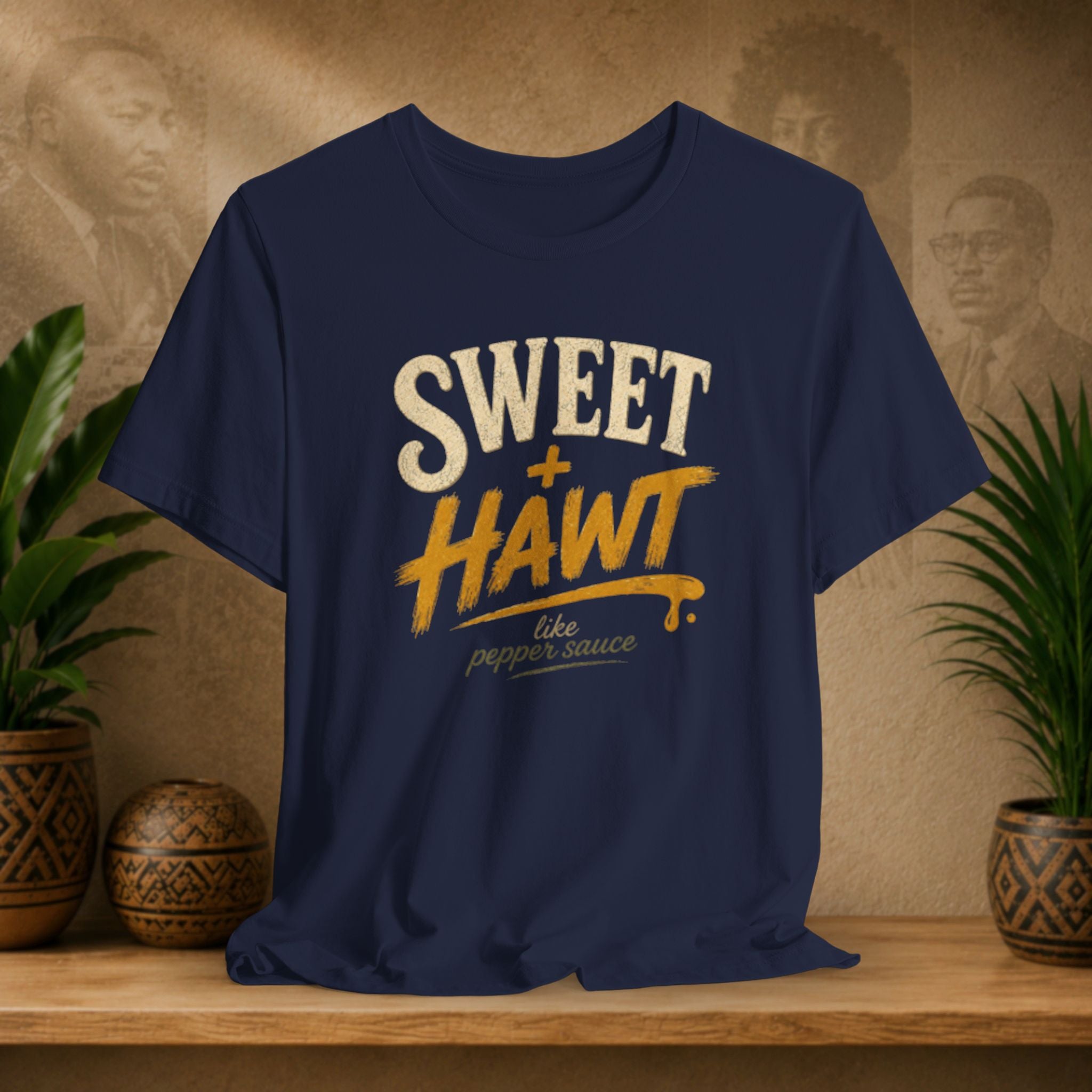 A navy blue graphic t-shirt displayed on a wooden shelf, flanked by potted plants and a textured background with sepia portraits. The shirt features a distressed graphic reading 'SWEET + HAWT like pepper sauce' in cream and yellow-orange lettering.
