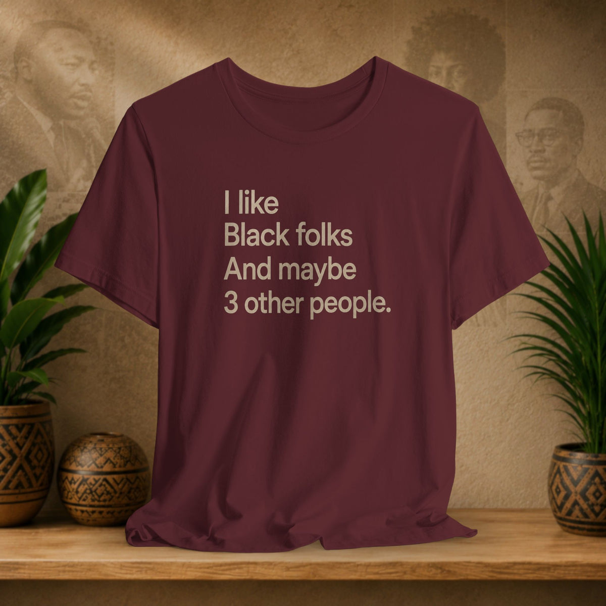 A maroon short-sleeve t-shirt with a beige printed quote on the front, displayed on a wooden shelf with potted plants and a textured background.