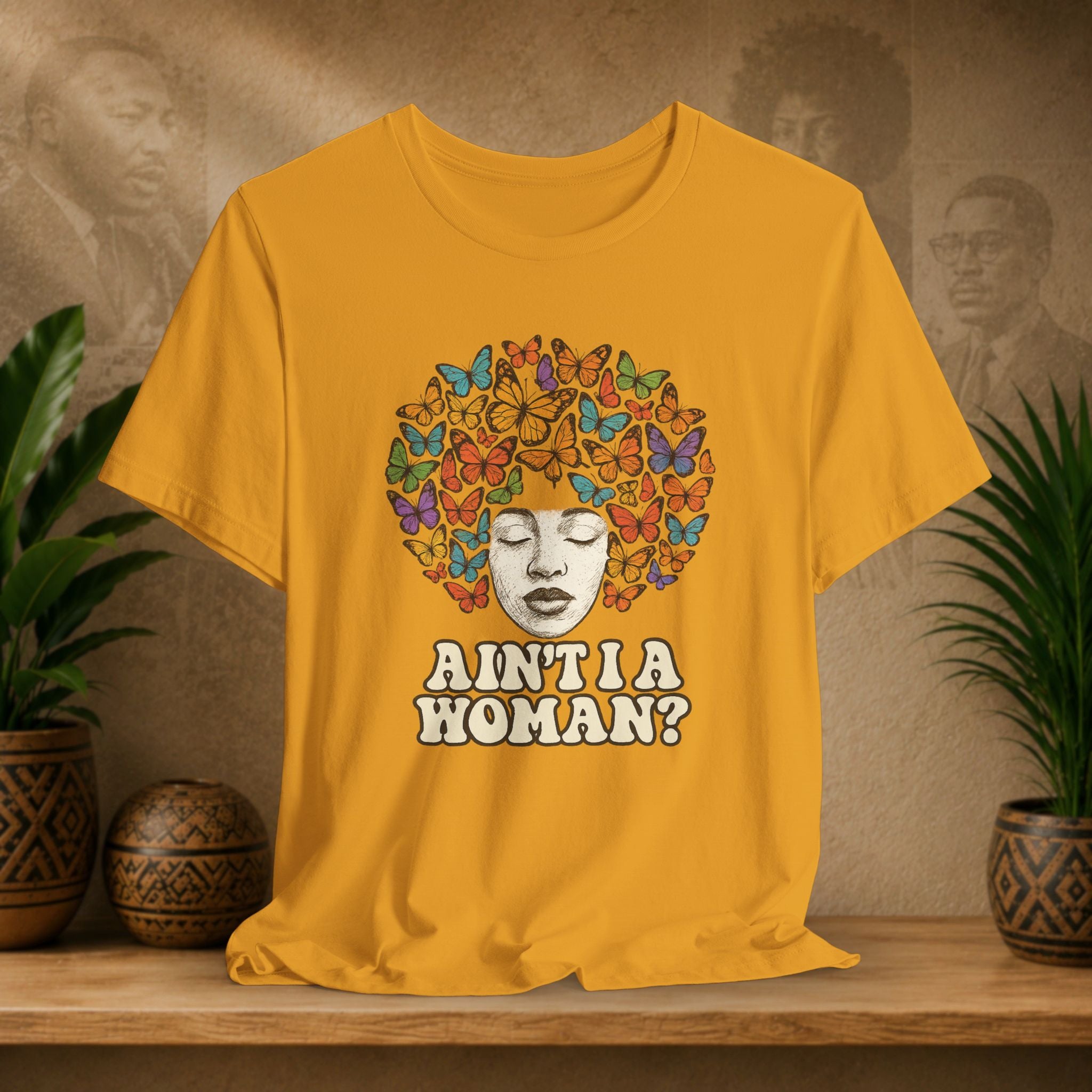 A mustard yellow graphic t-shirt displayed on a wooden shelf with potted plants in the background. The shirt features a graphic of a Black woman with a colorful afro hairstyle made of butterflies, with the text 'AIN'T I A WOMAN?' printed below.