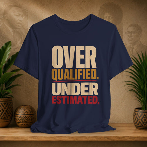 A navy blue graphic t-shirt displayed on a wooden shelf, flanked by potted green plants. The shirt features a bold text graphic reading 'OVER QUALIFIED.