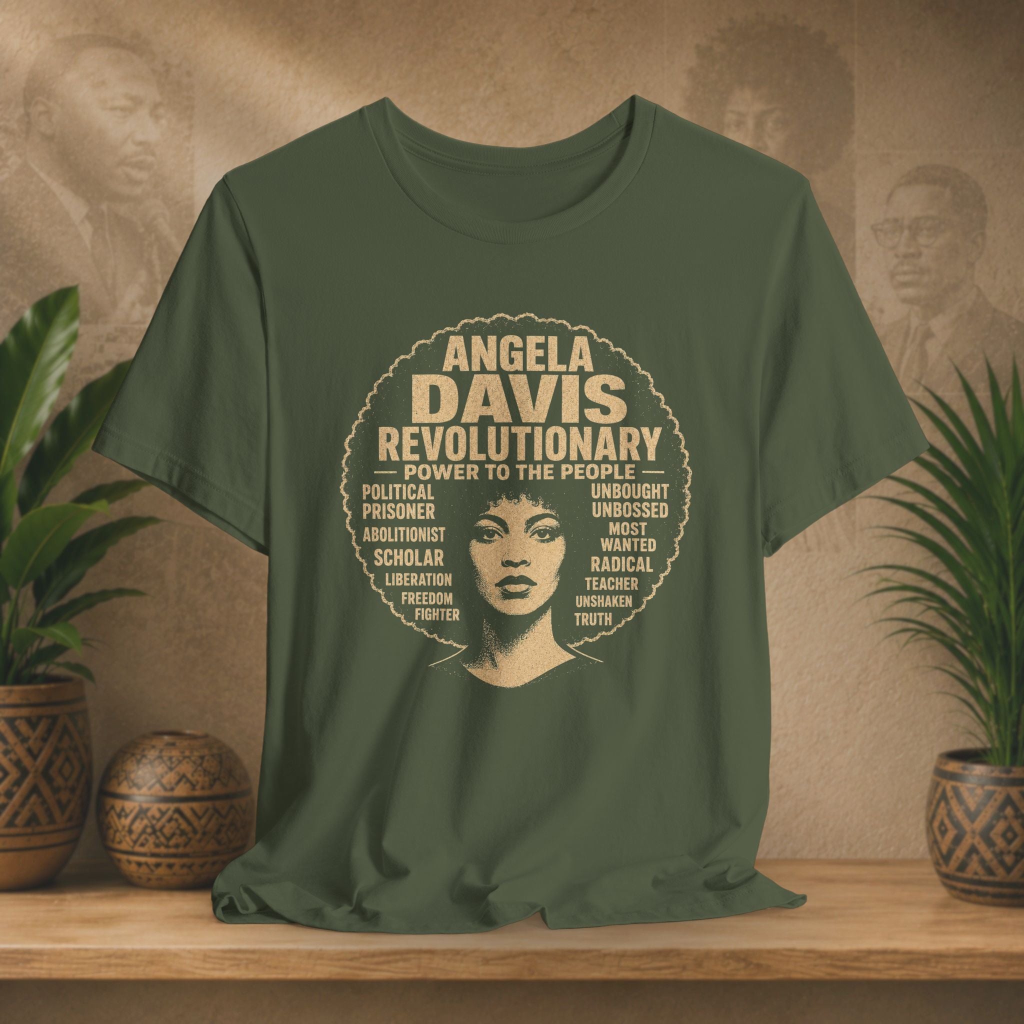 An olive green short-sleeve t-shirt with a vintage-style graphic of Angela Davis, displayed on a wooden shelf with potted plants in the background.