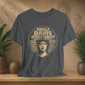 A dark gray graphic t-shirt featuring a vintage-style circular print of Angela Davis with her name and key revolutionary phrases in beige. The shirt is displayed on a wooden shelf in a styled setting with potted plants and a warm, textured background.