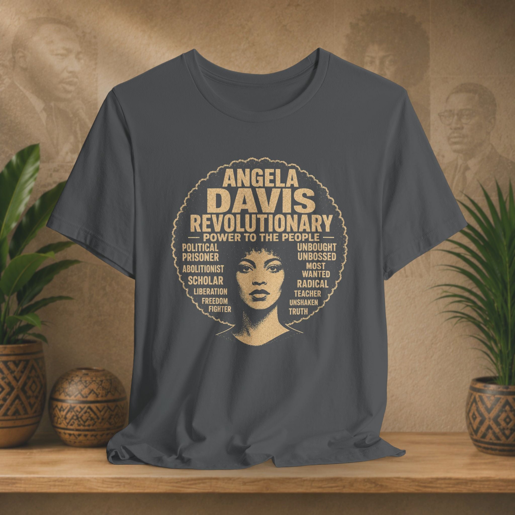 A dark gray graphic t-shirt featuring a vintage-style circular print of Angela Davis with her name and key revolutionary phrases in beige. The shirt is displayed on a wooden shelf in a styled setting with potted plants and a warm, textured background.