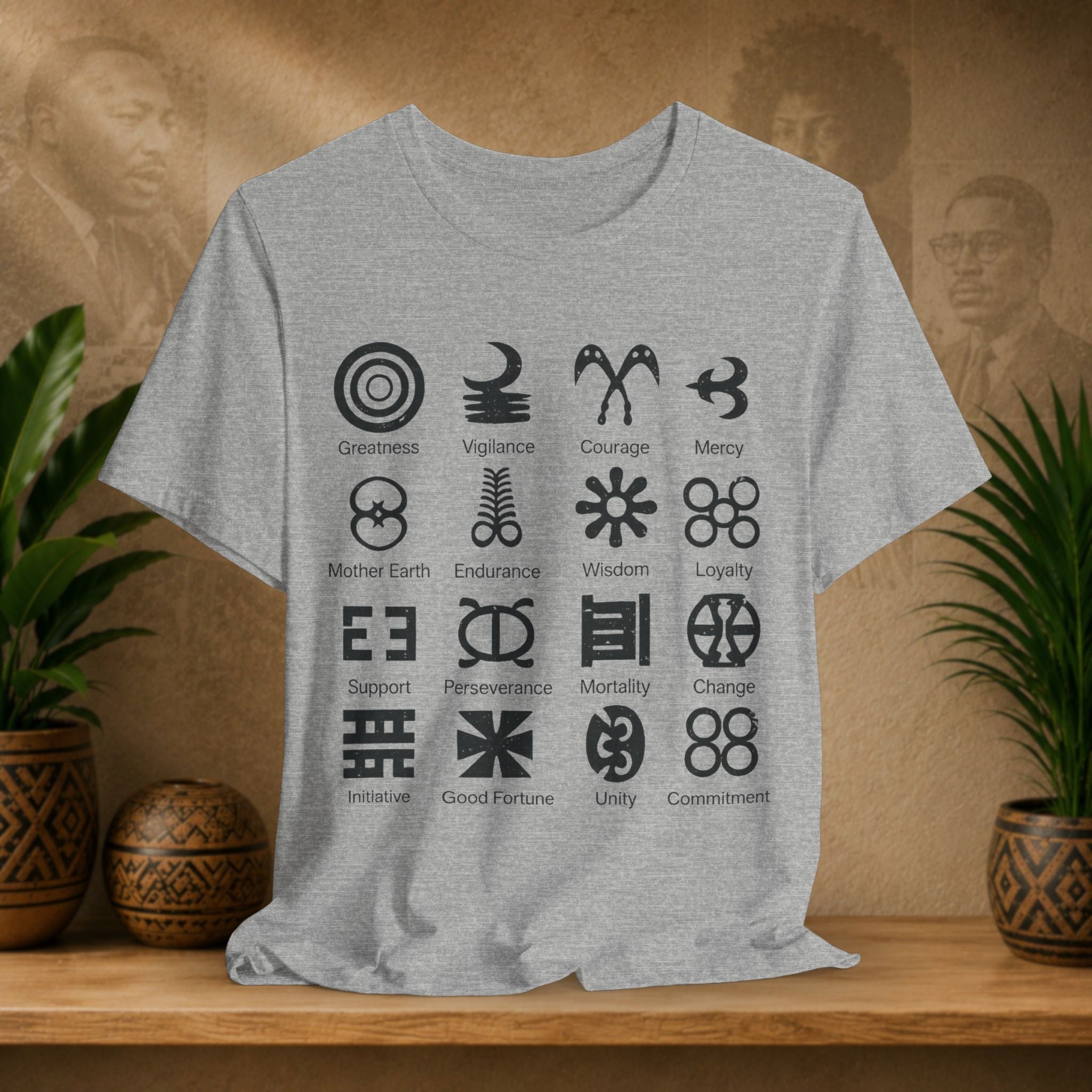 Grey t-shirt with black symbols and text on a wooden surface with plants and a textured wall in the background