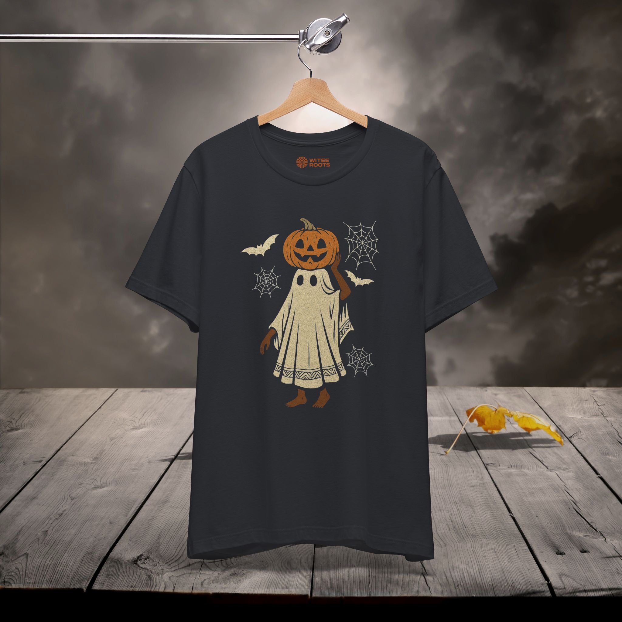 A black t-shirt hanging on a wooden hanger against a moody, smoky gray background with a rustic wooden plank floor. The t-shirt features a graphic of a cute cartoon pumpkin character dressed in a ghost costume, surrounded by bats, spiderwebs, and a small spider decoration on the floor.