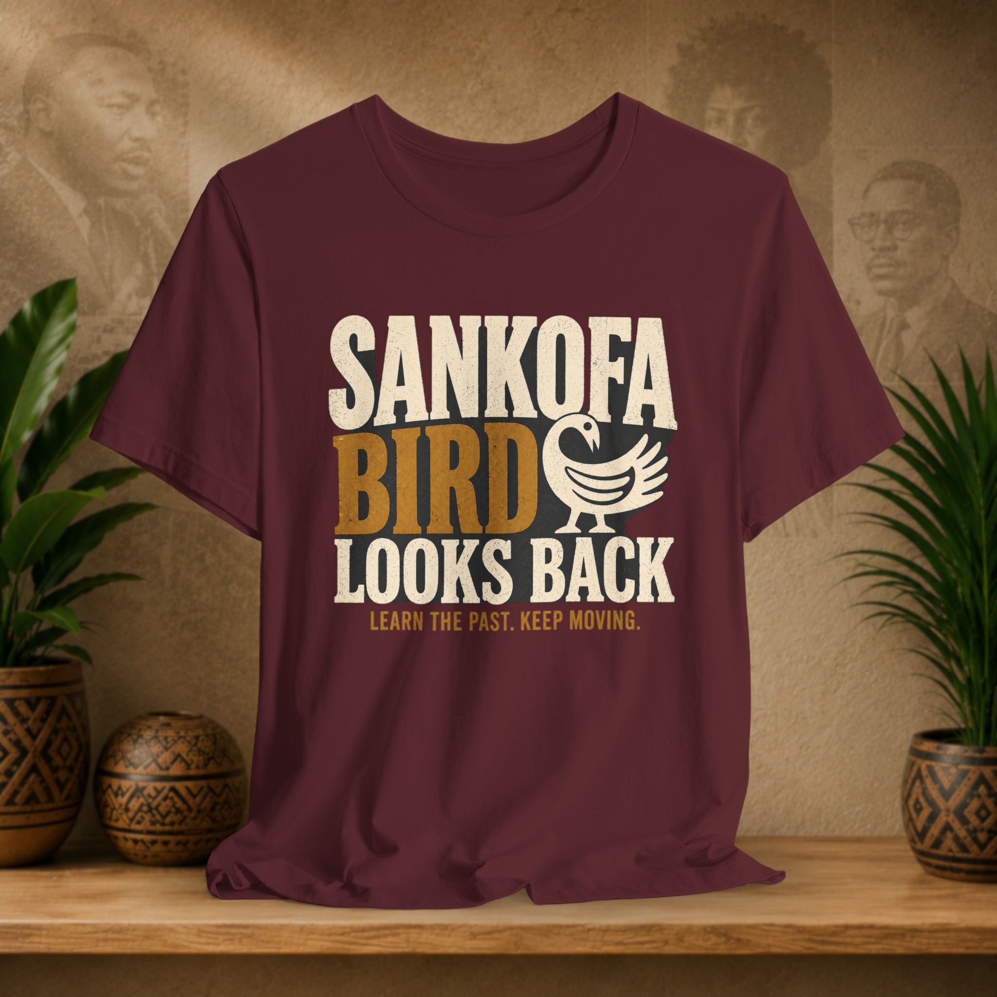 Maroon t-shirt with 'Sankofa Bird Looks Back' text on a wooden surface with plants and a textured wall background.