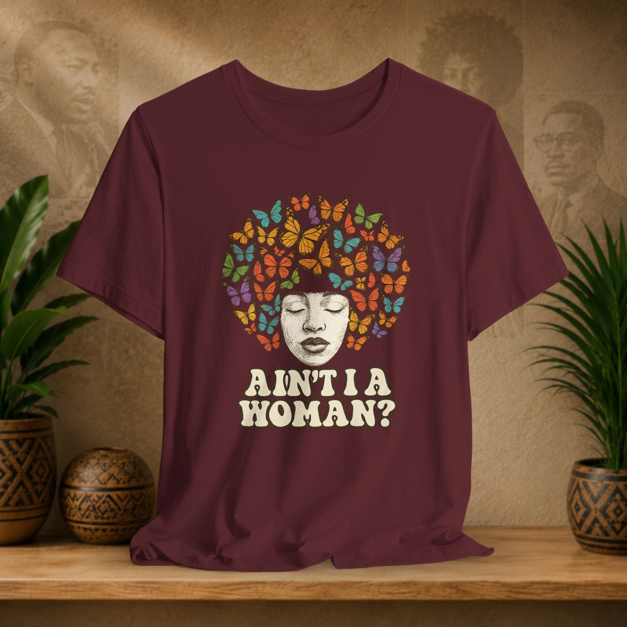 A maroon short-sleeve t-shirt displayed on a wooden shelf, featuring a graphic of a woman's face surrounded by colorful butterflies and the text 'AIN'T I A WOMAN?'. The background shows a warm textured wall with faded portraits and potted plants.