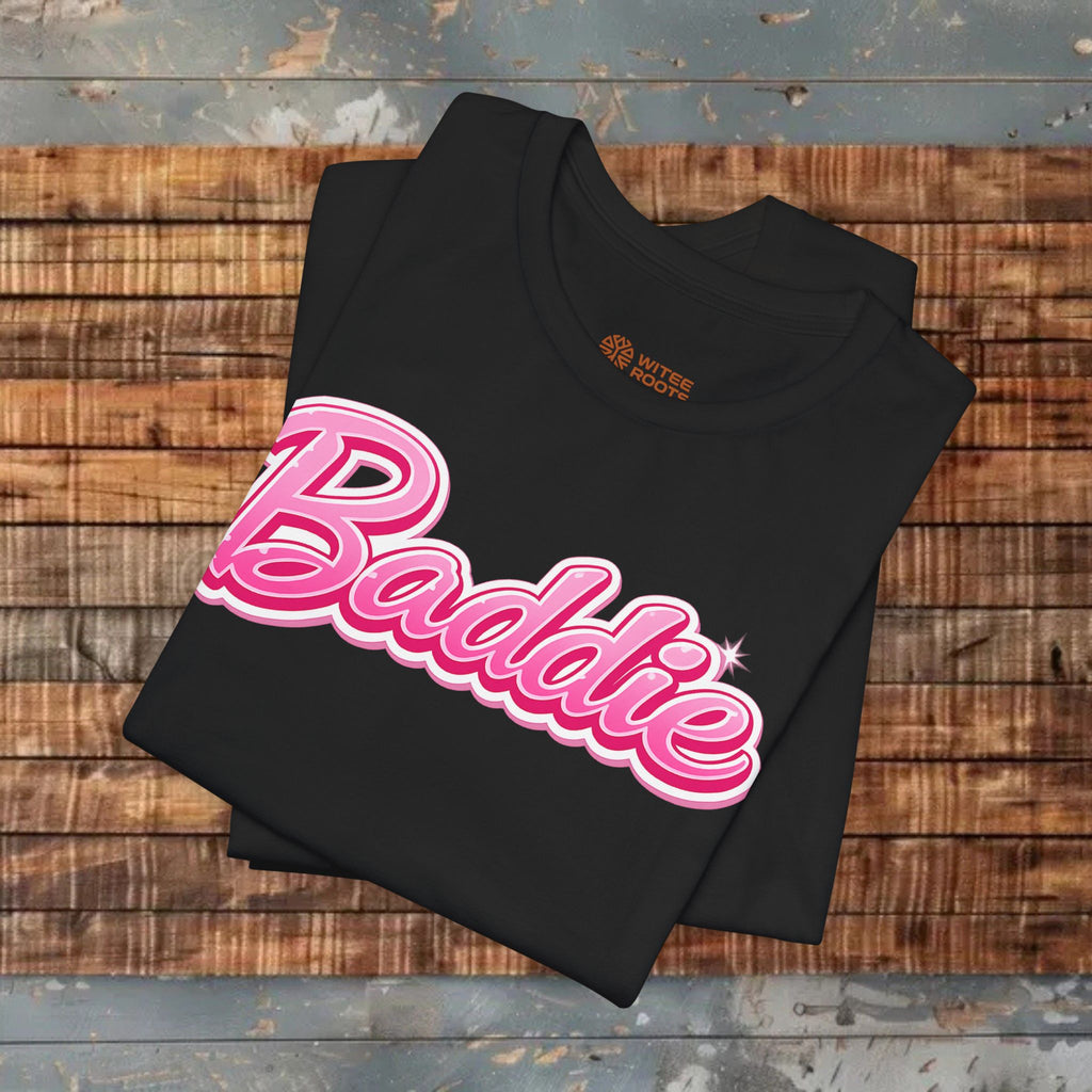 A folded black t-shirt with a pink and white 'Badbie' graphic across the front, displayed on a rustic wooden plank surface.