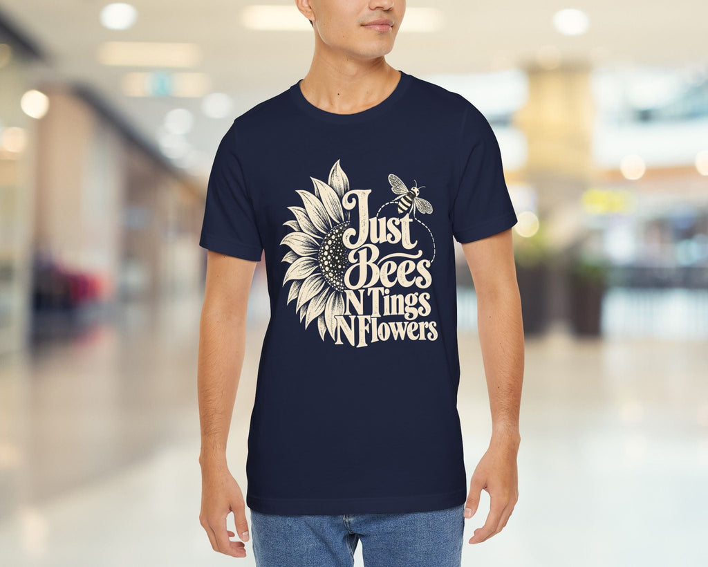 A man wearing a navy blue t-shirt with a white graphic that reads 'Just Bees N' Things Flowers' alongside a sunflower and bee illustration, standing in a brightly lit, blurred indoor hallway.