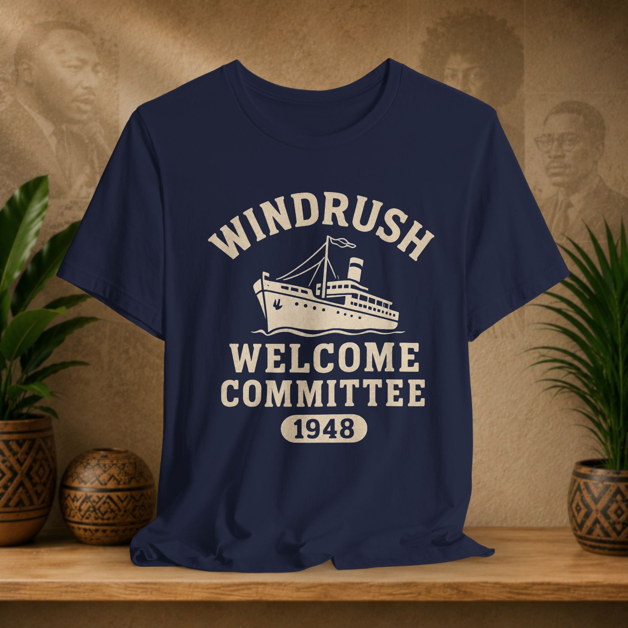 Navy blue t-shirt with 'Windrush Welcome Committee 1948' design on a wooden surface with plants and a vintage-style background.