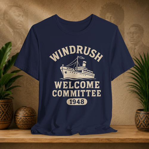Navy blue t-shirt with 'Windrush Welcome Committee 1948' design on a wooden surface with plants and a vintage-style background.