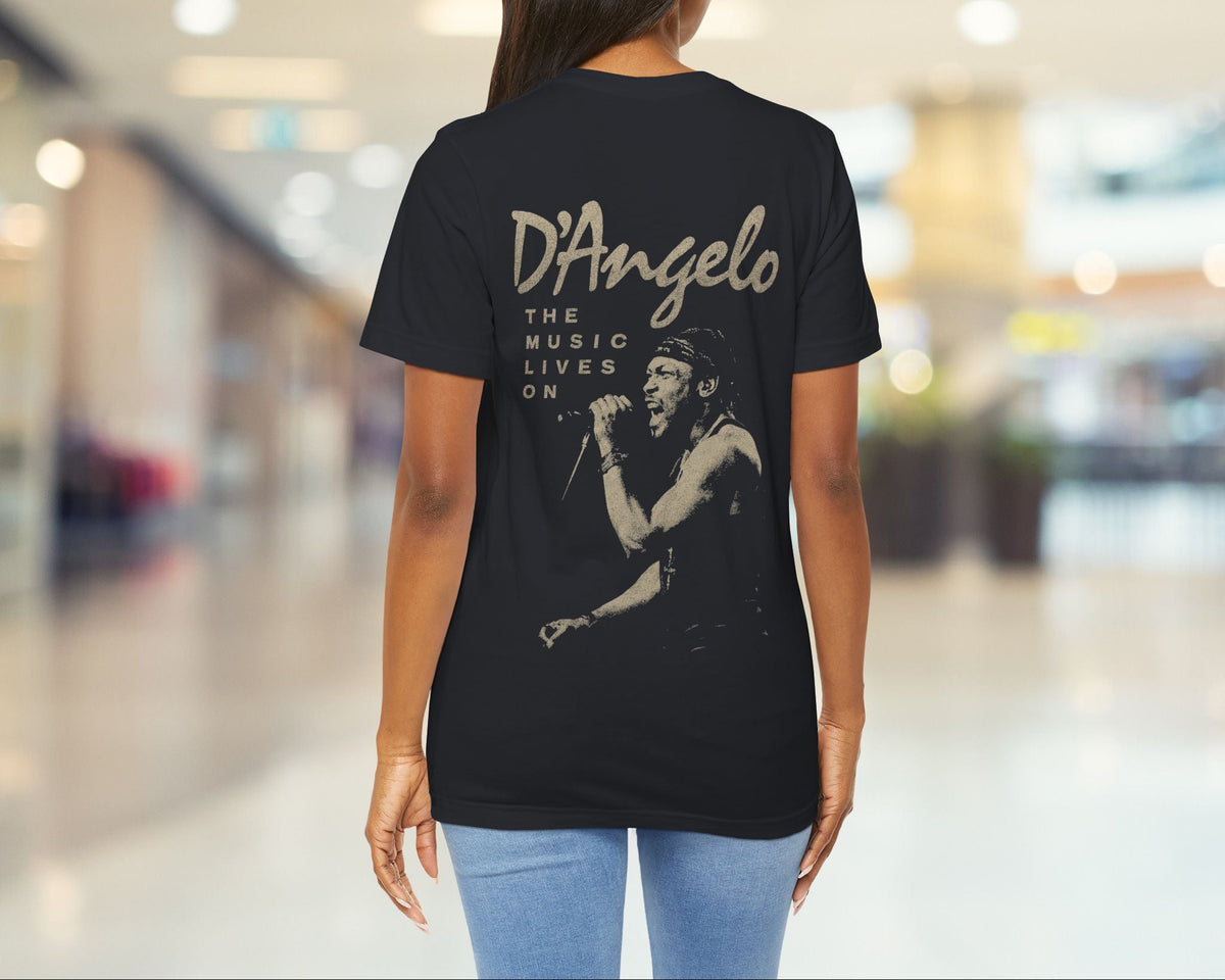 A woman viewed from behind wearing a black t-shirt with a large beige graphic print of D'Angelo on the back, set against a blurred indoor hallway background.