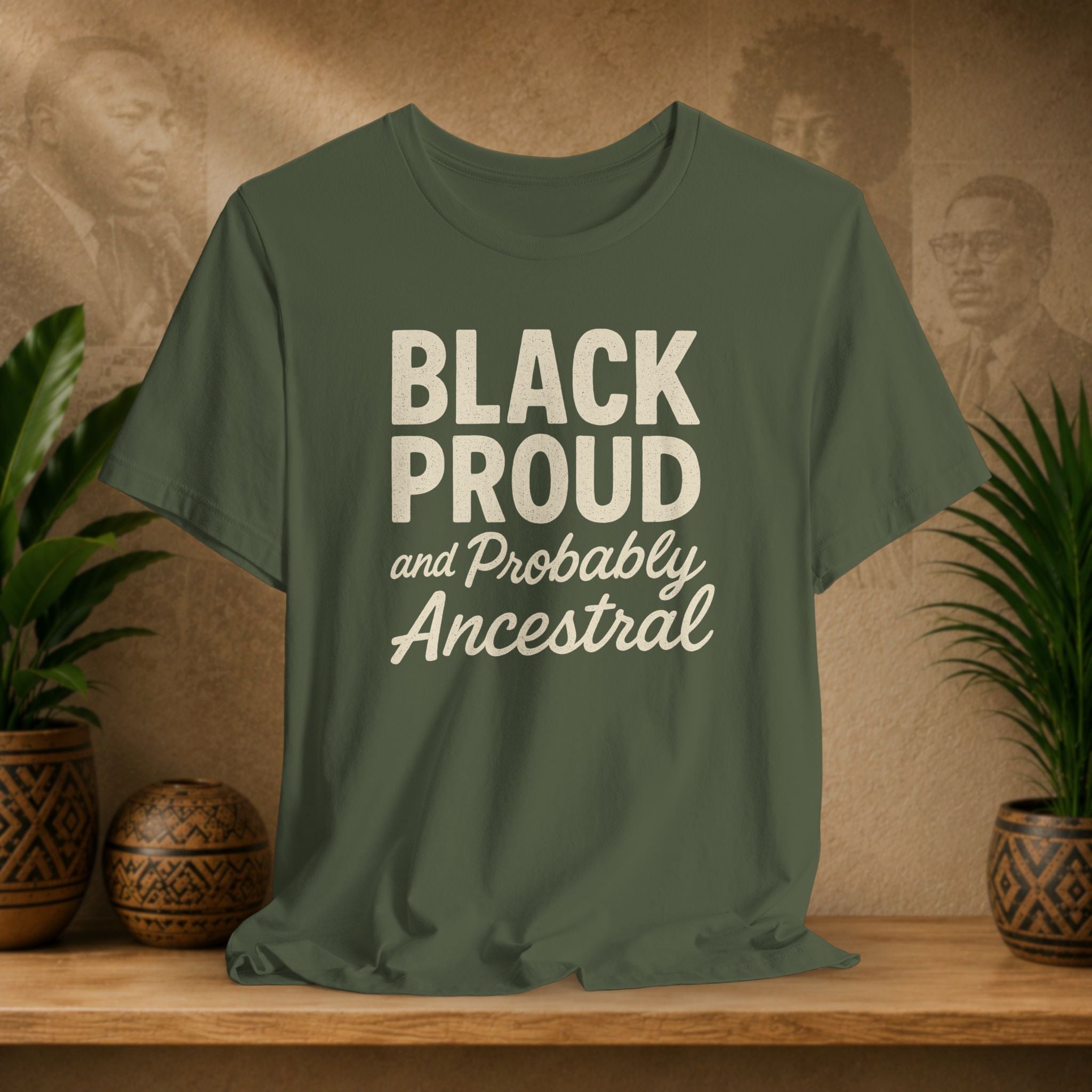 Green t-shirt with 'Black Proud and Probably Ancestral' text on a wooden surface with plants and a decorative wall in the background.