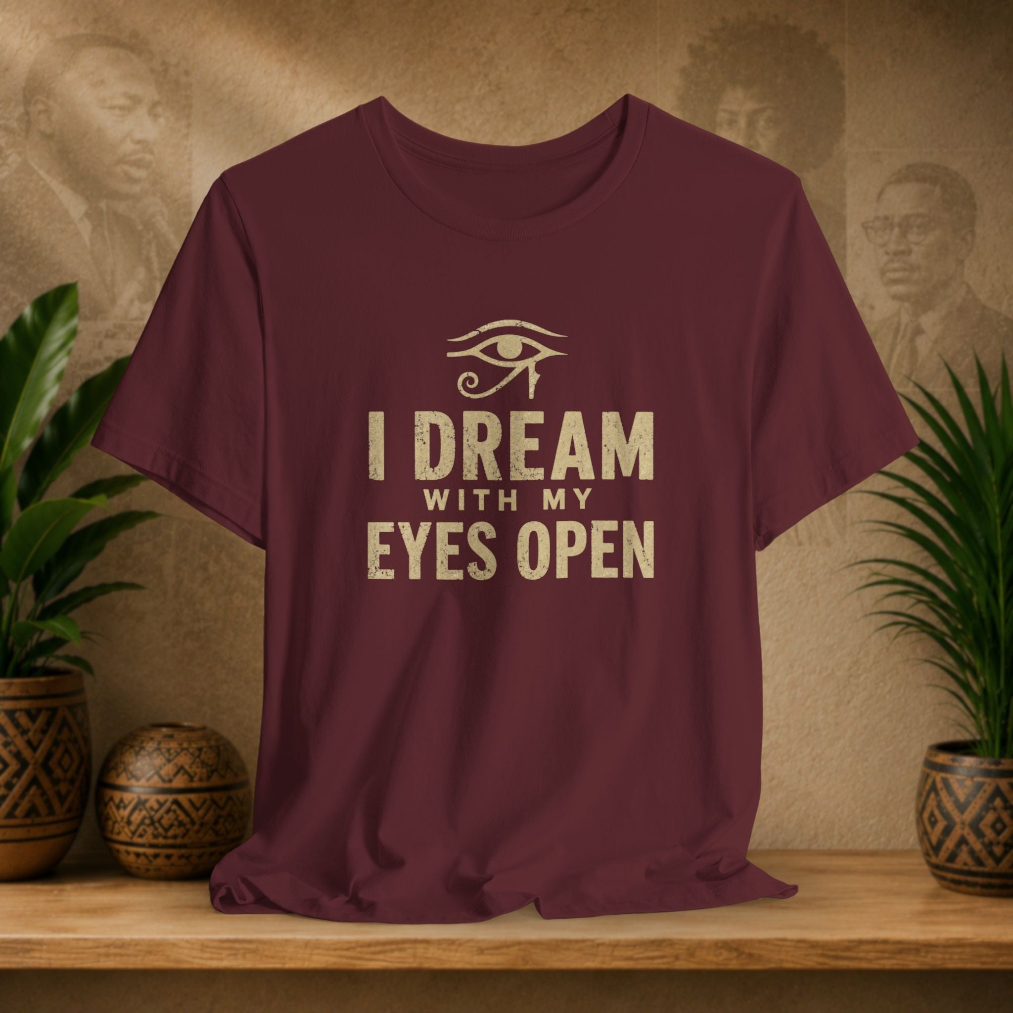 Maroon t-shirt with 'I Dream with My Eyes Open' text and eye symbol on a wooden surface with plants and wall art.