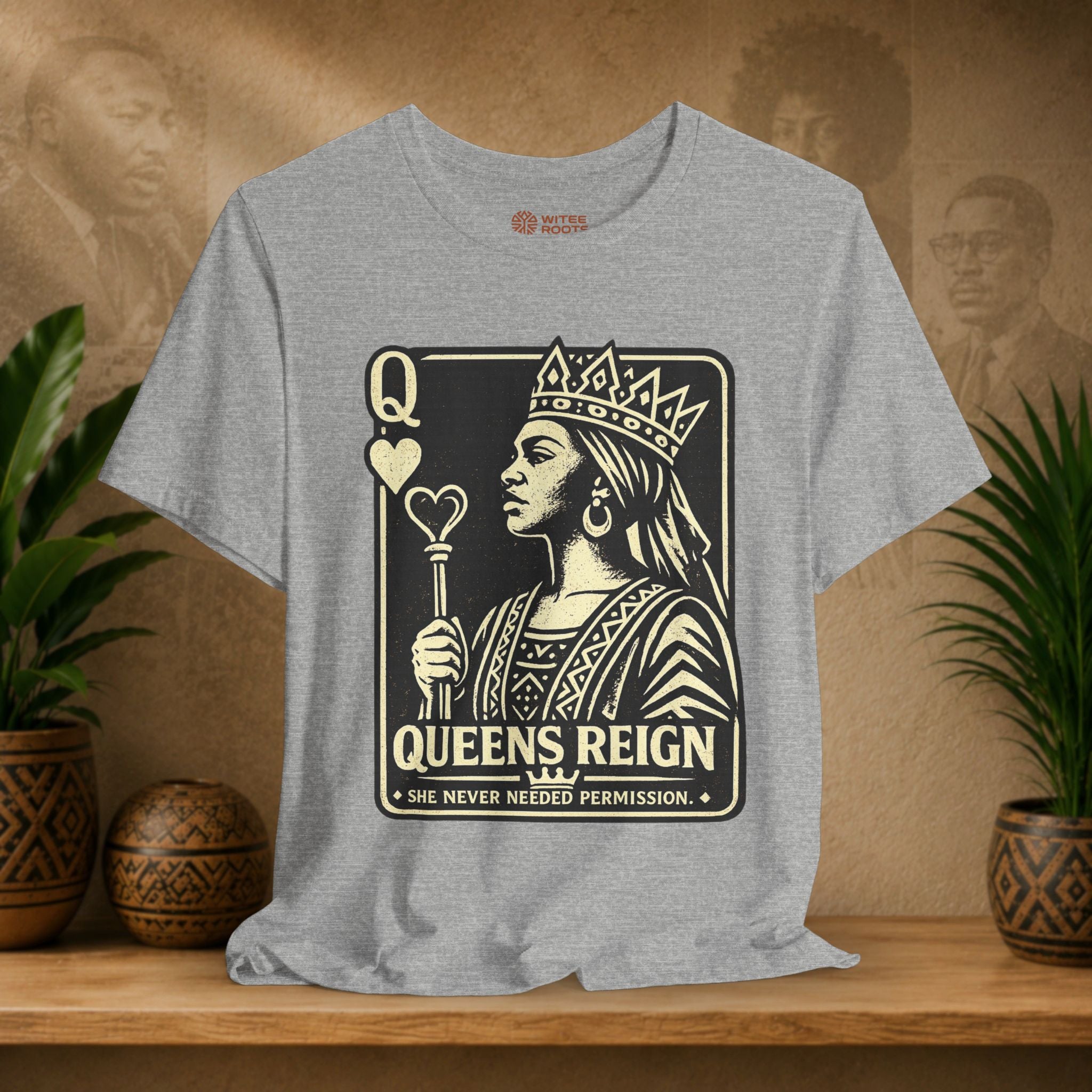 Gray t-shirt with 'Queens Reign' graphic design on a wooden shelf with plants and a textured wall background.